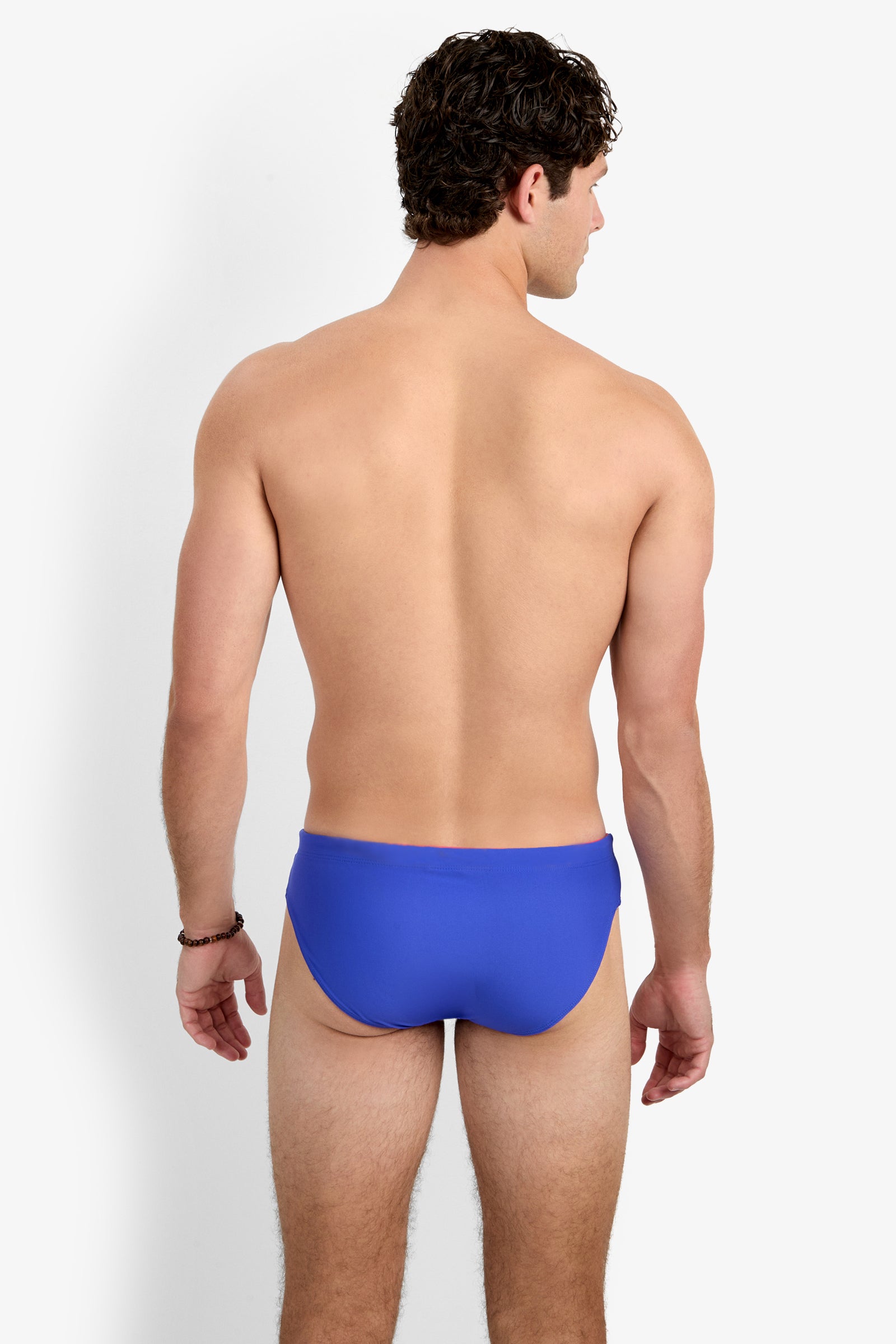 A man with short curly hair is seen from behind, wearing the bright blue Palm Beach Swim Brief. He stands against a plain white background with his arms relaxed by his sides.