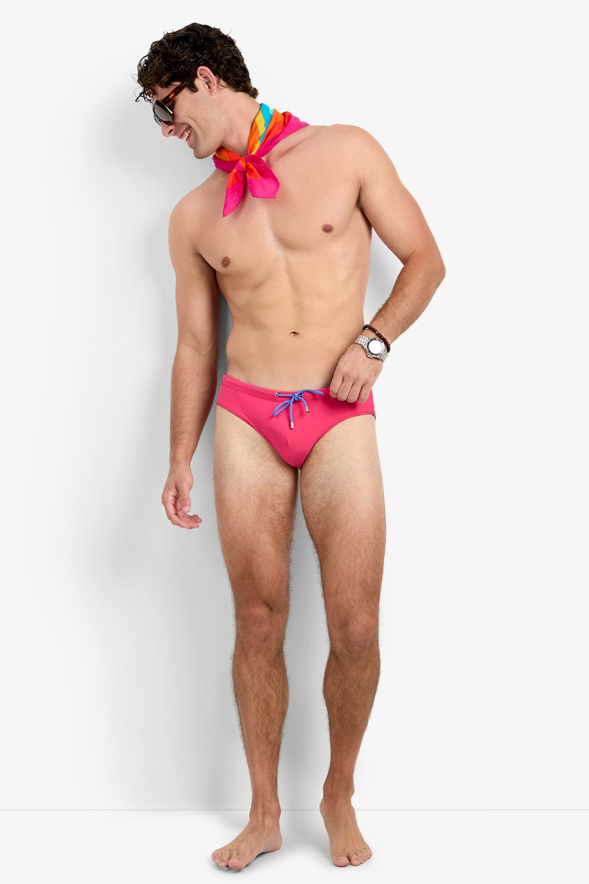 A smiling man in the PALM BEACH SWIM BRIEF stands barefoot against a plain white background, wearing sunglasses, a colorful scarf, and a watch, looking to his left.