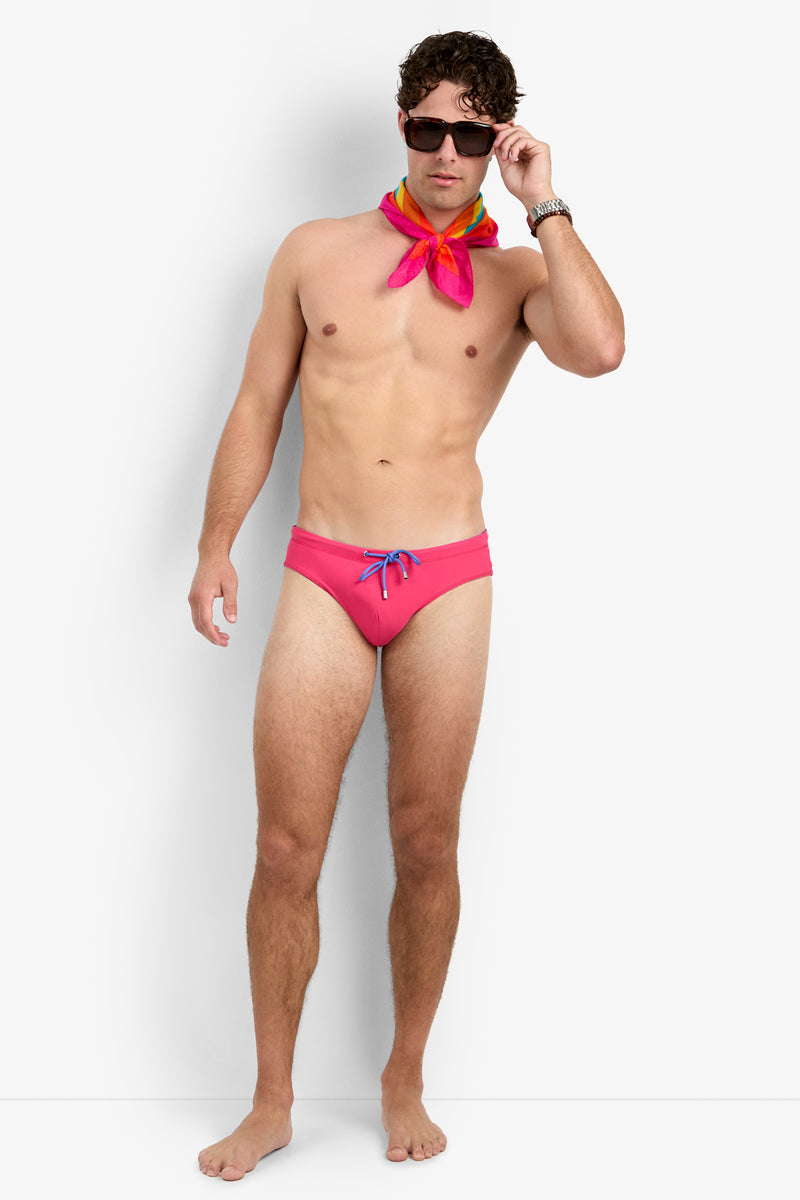 A man stands barefoot against a plain background, wearing the PALM BEACH SWIM BRIEF in pink, a colorful scarf around his neck, dark sunglasses, and a wristwatch, with one hand adjusting his sunglasses.