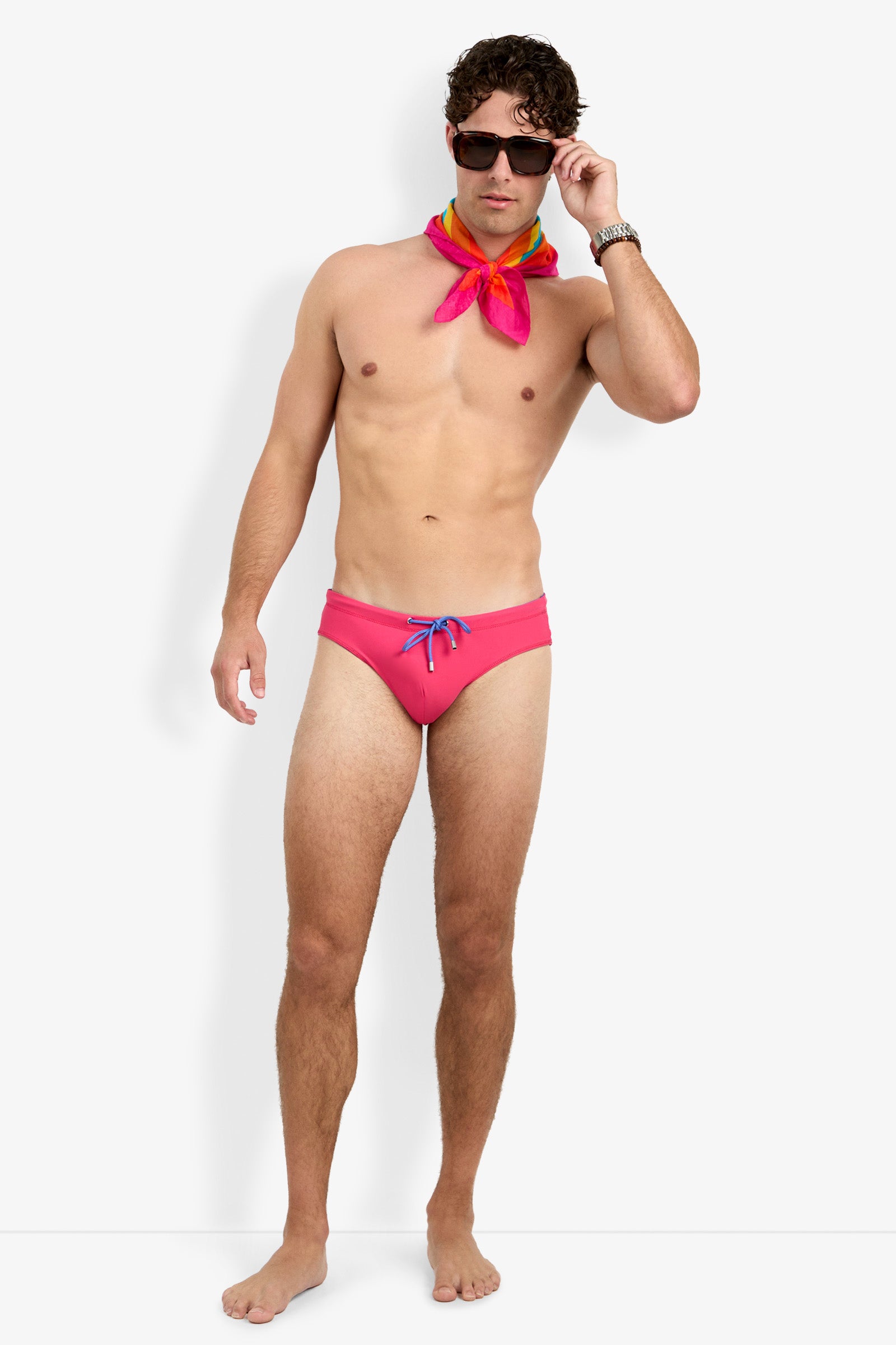 A man stands barefoot against a white background, wearing the Palm Beach Swim Brief with a blue drawstring, a colorful neck scarf, dark sunglasses, and a wristwatch, looking forward while adjusting his sunglasses.