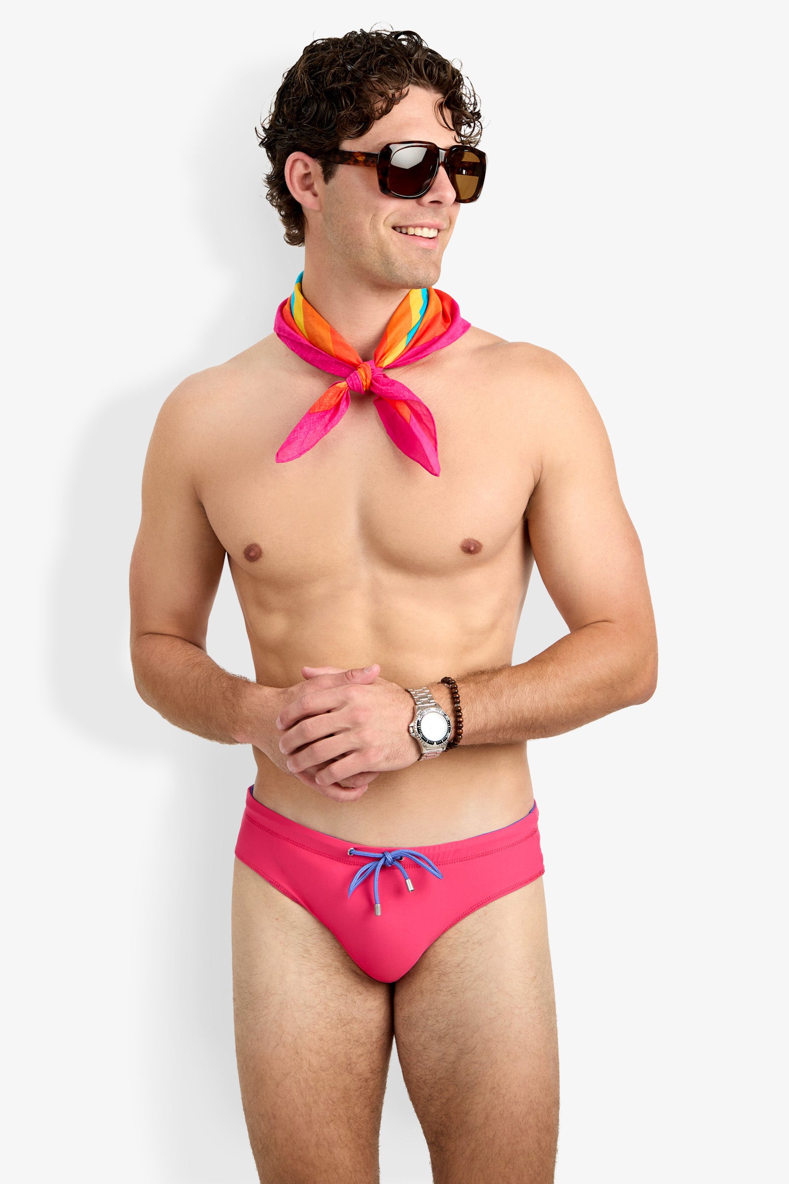 A smiling, shirtless man in dark sunglasses, a colorful neck scarf, wristwatch, and bright pink Palm Beach Swim Brief poses against a plain white background.