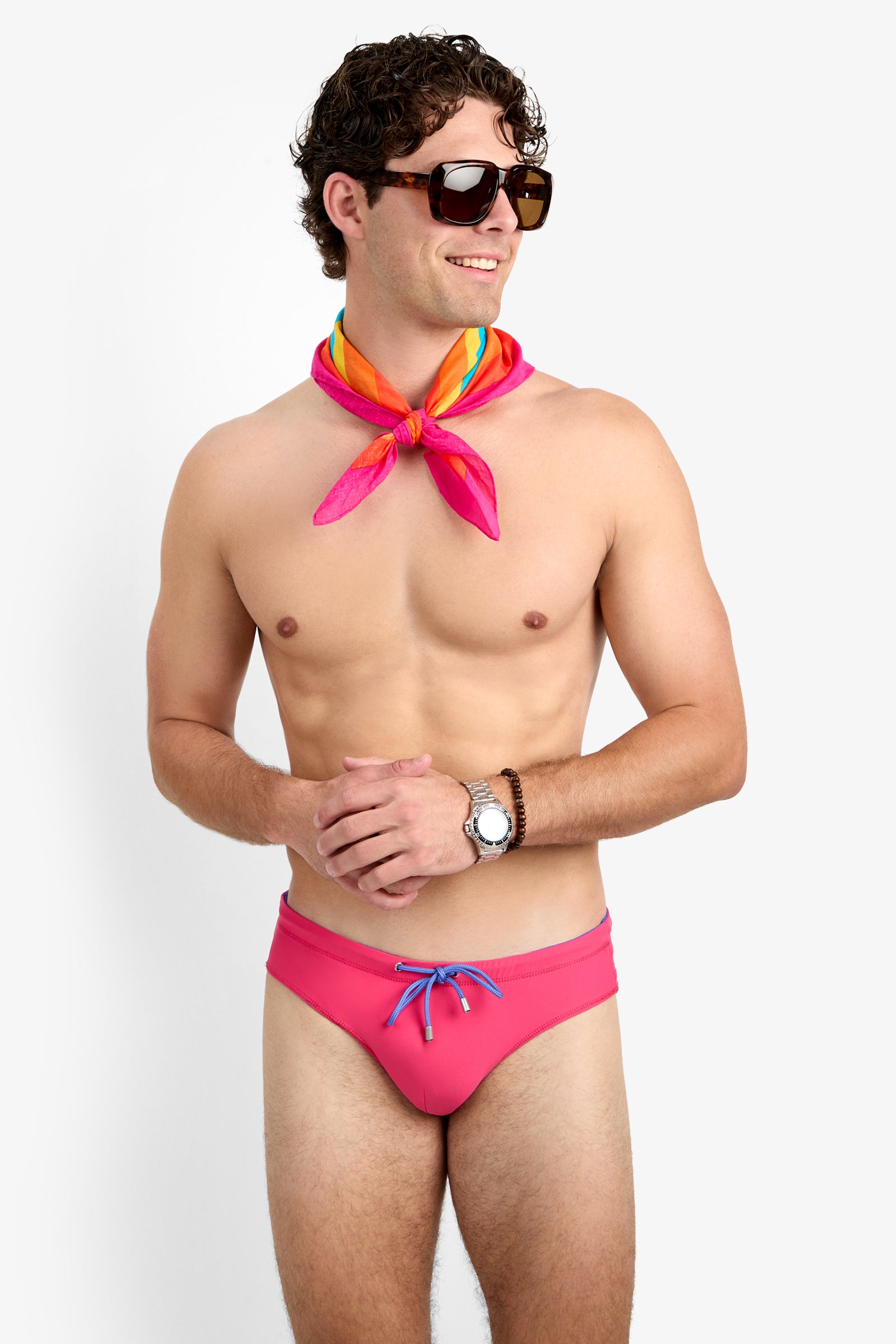 A smiling man in dark sunglasses, a colorful scarf, a wristwatch, and Palm Beach Swim Briefs stands against a plain white background with his hands loosely clasped.