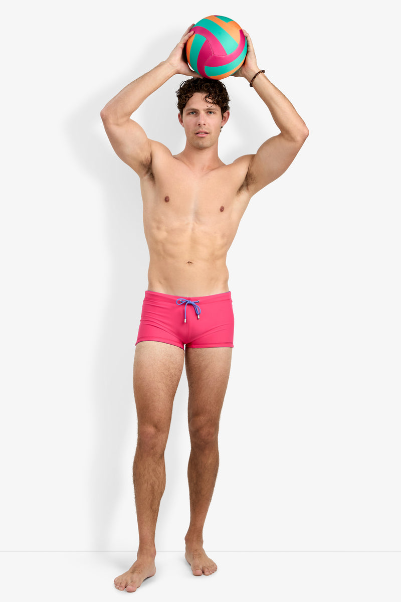 A man wearing Seaside Beach Swim Trunk stands barefoot, holding a colorful volleyball above his head against a plain white background.