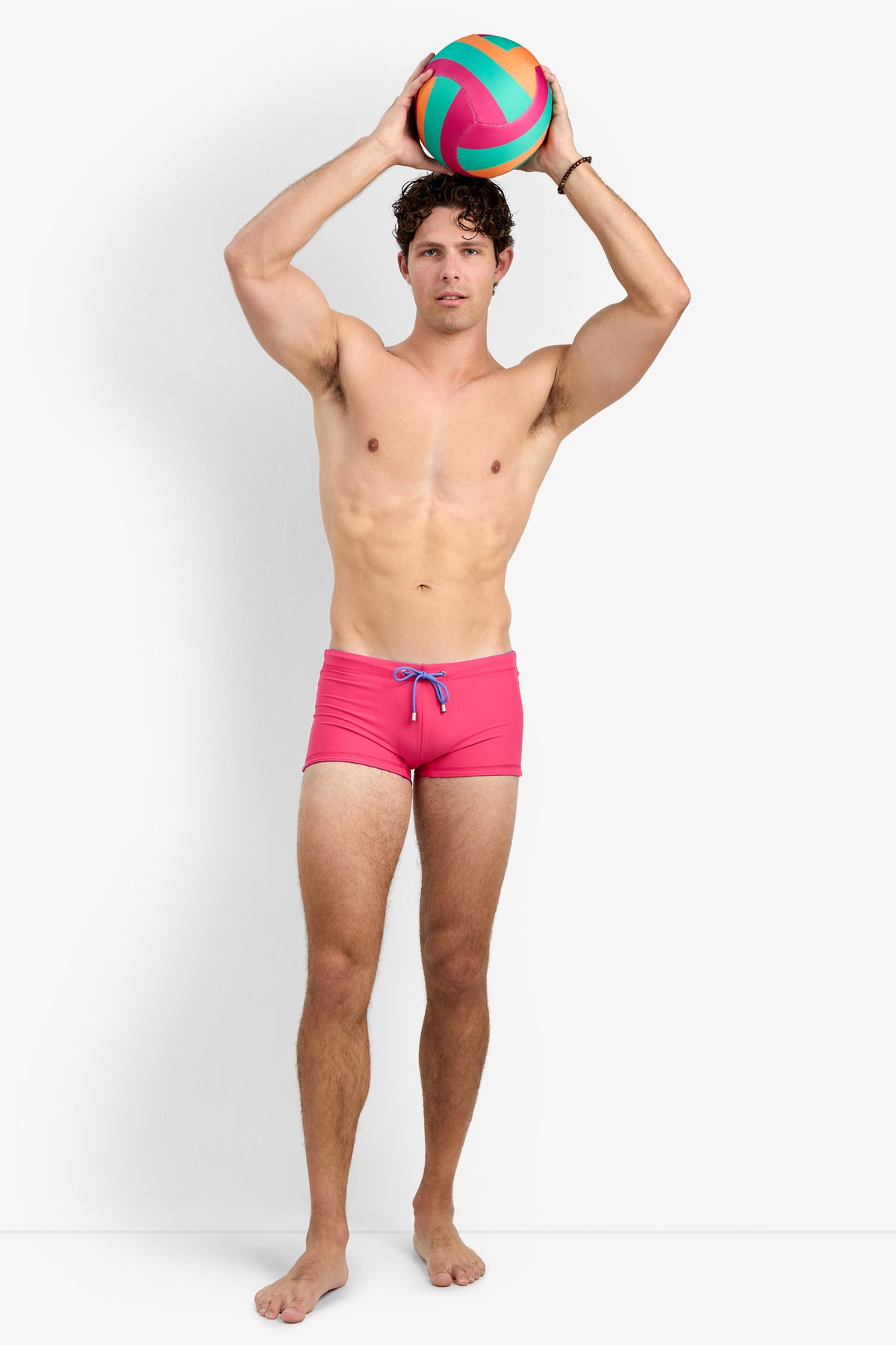 A man stands barefoot in Seaside Beach Swim Trunk, holding a colorful beach ball over his head against a plain white background.
