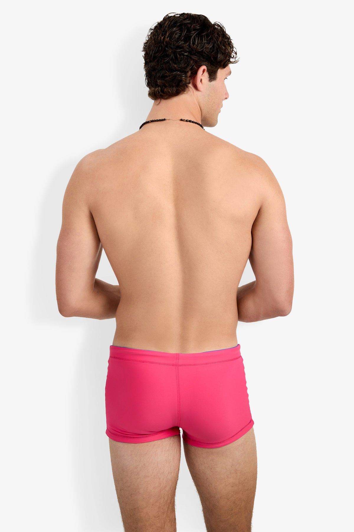 A man with short curly hair stands with his back to the camera, wearing Seaside Beach Swim Trunk in bright pink against a plain white background.