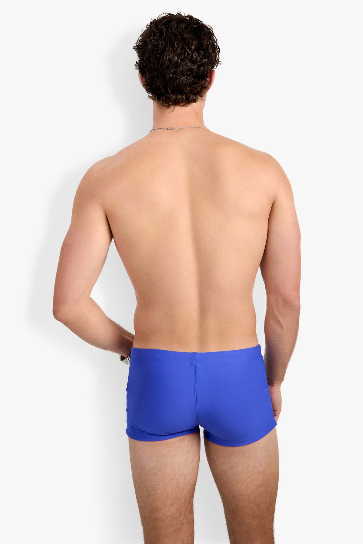 A shirtless man with curly brown hair stands with his back to the camera, wearing Seaside Beach Swim Trunk in bright blue against a plain white background.