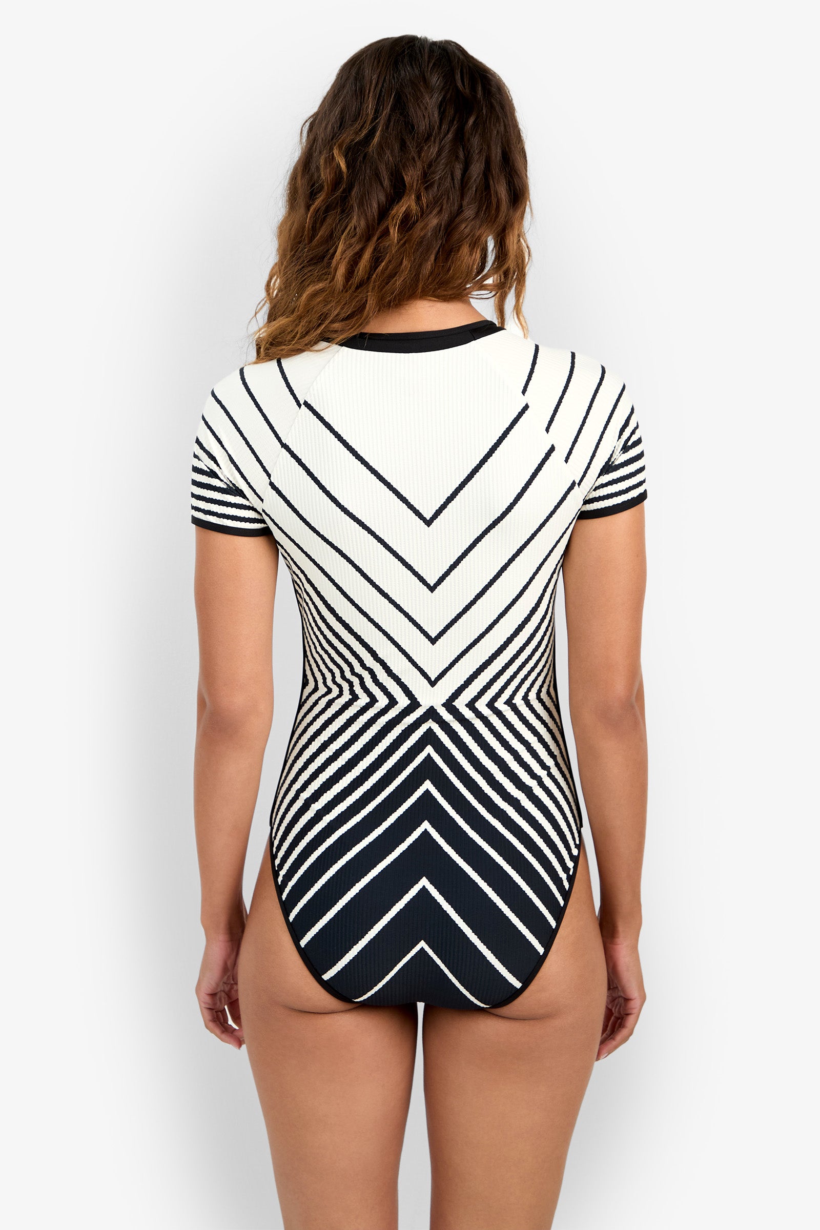 A woman faces away, wearing the Short-Sleeve Zip-Up Paddlesuit—black and white with bold geometric stripes in a V-shape. The background is plain white.