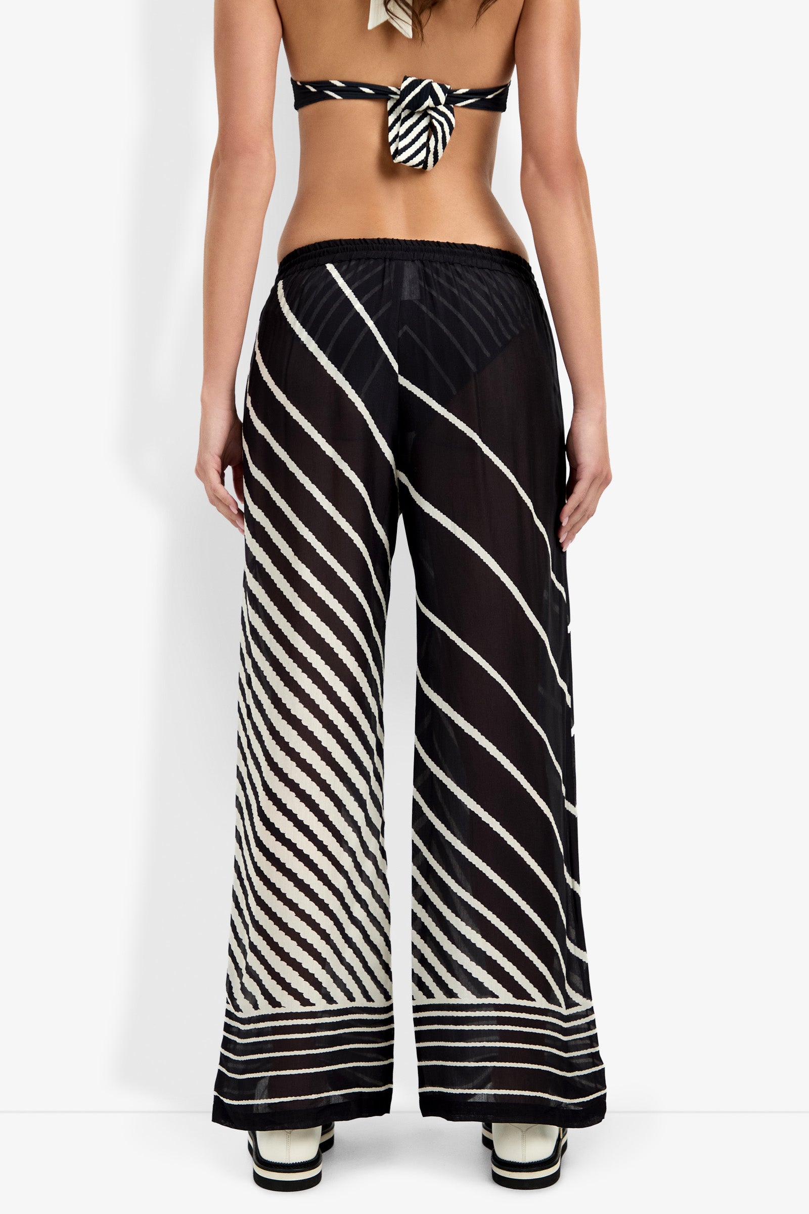 A woman, back turned, wears the Net Stripe Beach Pant—sheer black wide-leg pants with diagonal white stripes—paired with a matching bikini top and platform sandals; her back is exposed.