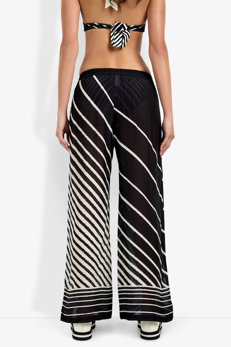 A woman, back turned, wears the Net Stripe Beach Pant—sheer black wide-leg pants with diagonal white stripes—paired with a matching bikini top and platform sandals; her back is exposed.