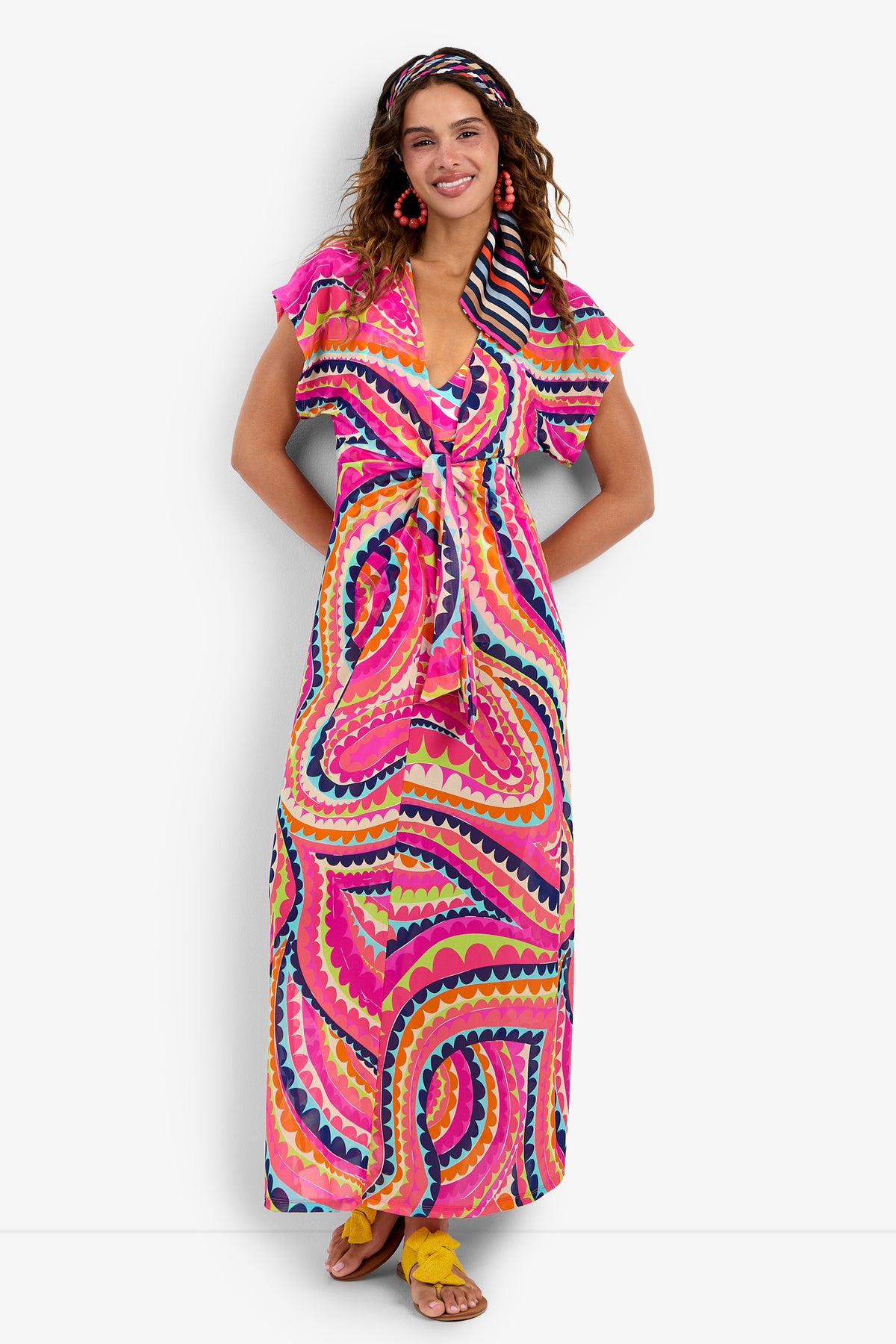 A woman smiles against a white background, wearing the Cover-Up Maxi Dress featuring a long, colorful pattern in pink, orange, yellow, and blue with a matching headband, paired with yellow sandals and pink earrings.