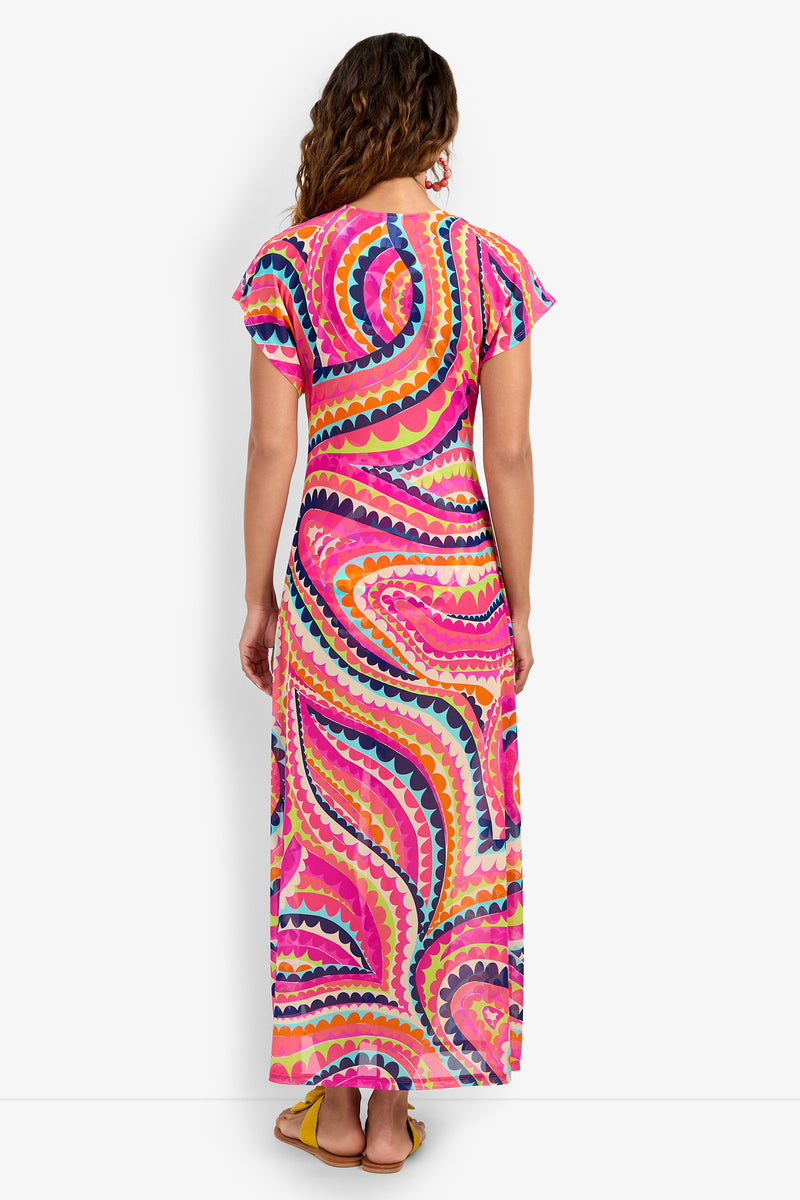 A person with wavy brown hair faces away, modeling the Cover-Up Maxi Dress featuring colorful pink, yellow, blue, and orange swirls, paired with yellow sandals and set against a white background.