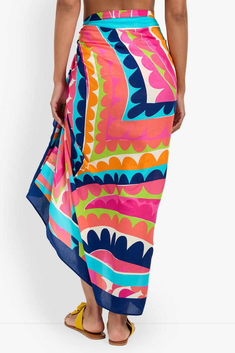 A person stands facing away from the camera against a white background, wearing the Pareo—a colorful wrap skirt with pink, orange, blue, and yellow geometric shapes—paired with yellow sandals.