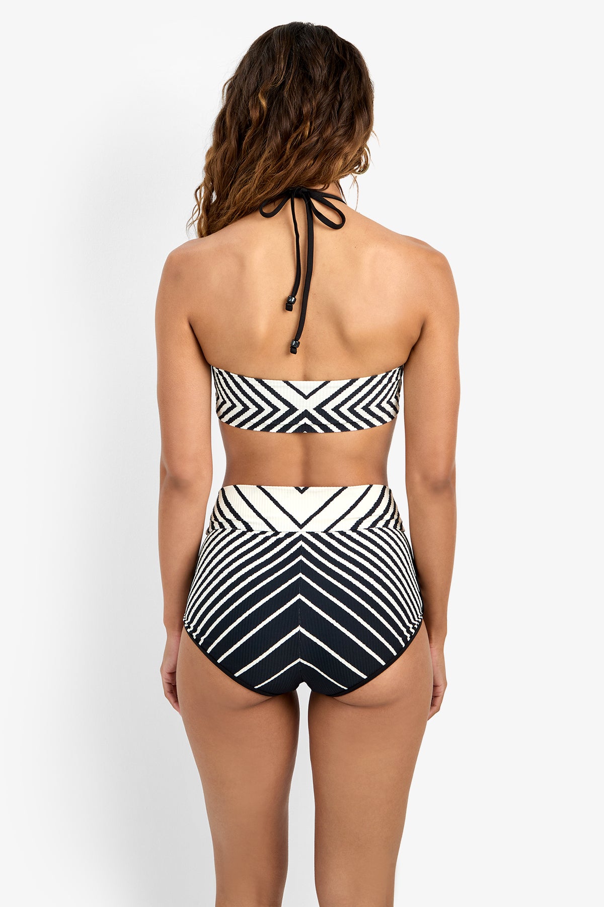 Woman with long wavy hair stands against a white background, facing away and wearing the Net Stripe High Neck Bikini Top in black and white with a geometric pattern and high-waisted bottoms.