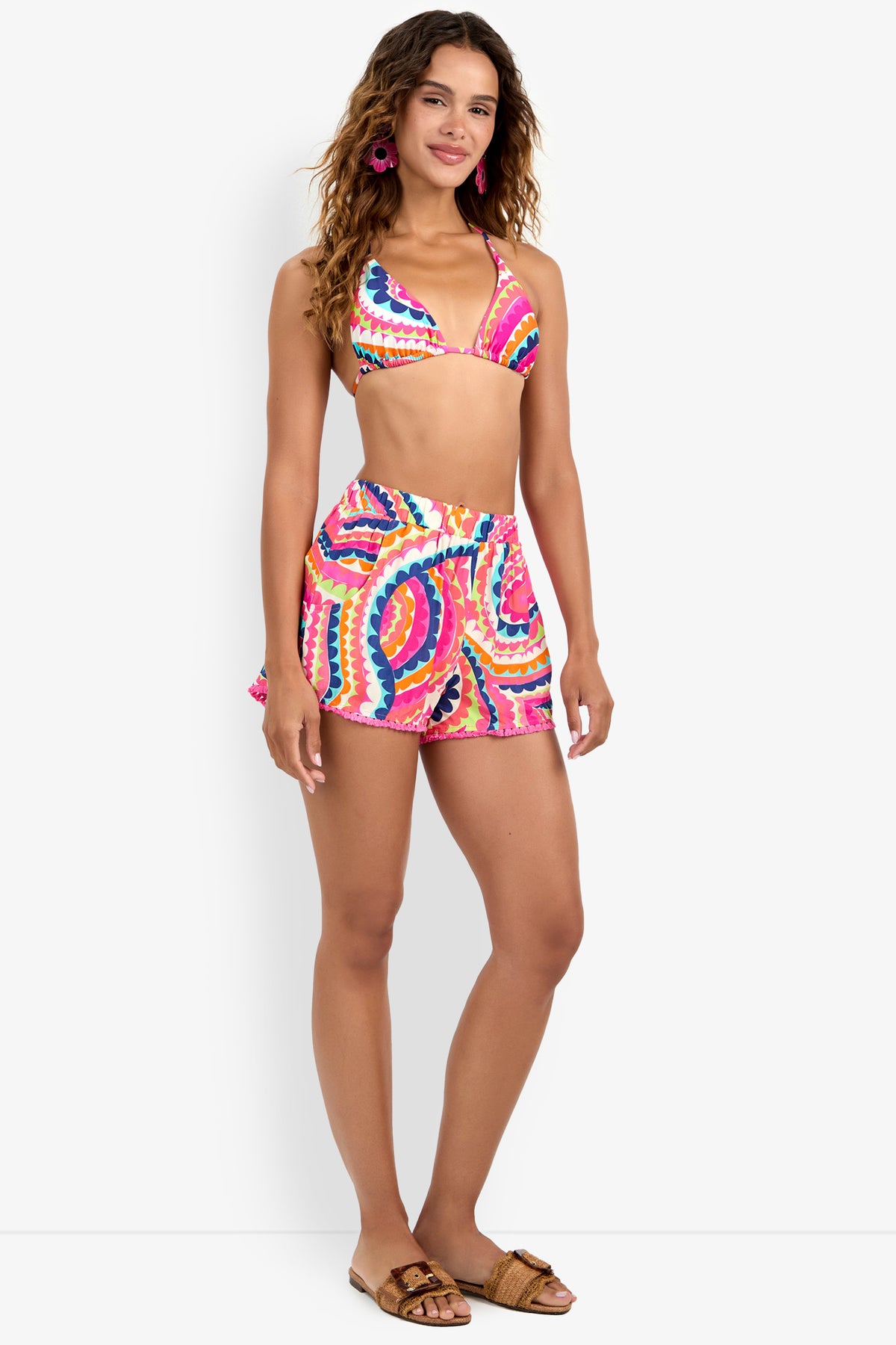 A woman smiles against a plain white background, wearing a Triangle Bikini Top and matching shorts with a colorful tie-dye pattern. She has long, wavy hair, pink earrings, and brown sandals.