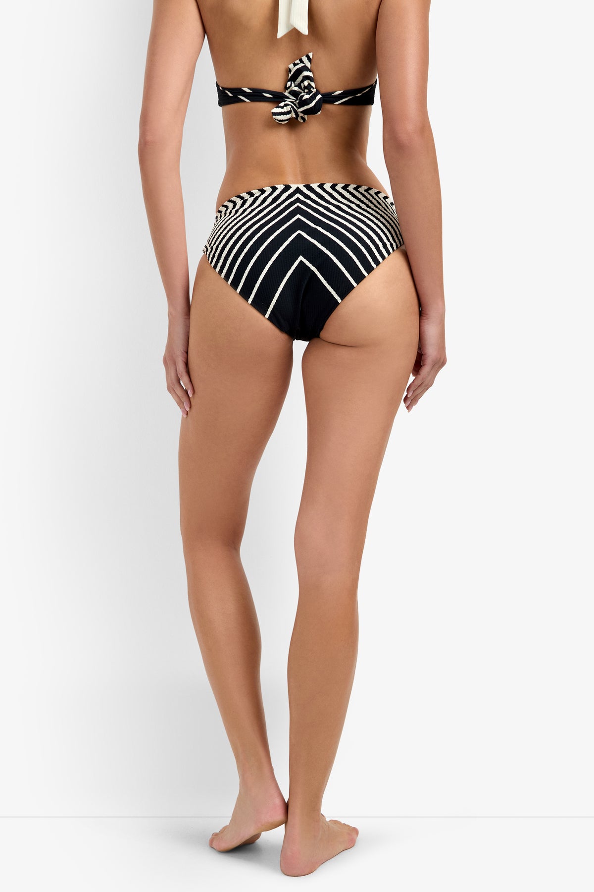 A woman stands barefoot, back-facing, wearing the Net Stripe Hipster Bikini Bottom with a black and white chevron pattern. The plain white background highlights the swimwear’s bold stripes and knot detail.