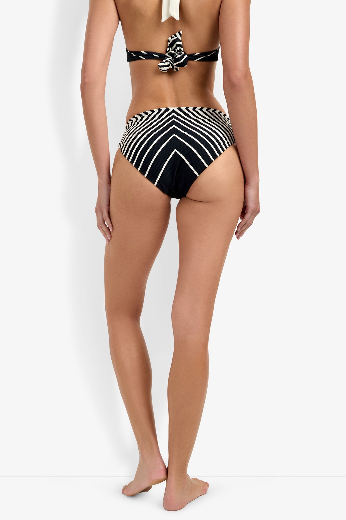 A woman stands barefoot facing away, wearing the Net Stripe Hipster Bikini Bottom in black and white stripes with downward-pointing chevrons. The background is plain white.