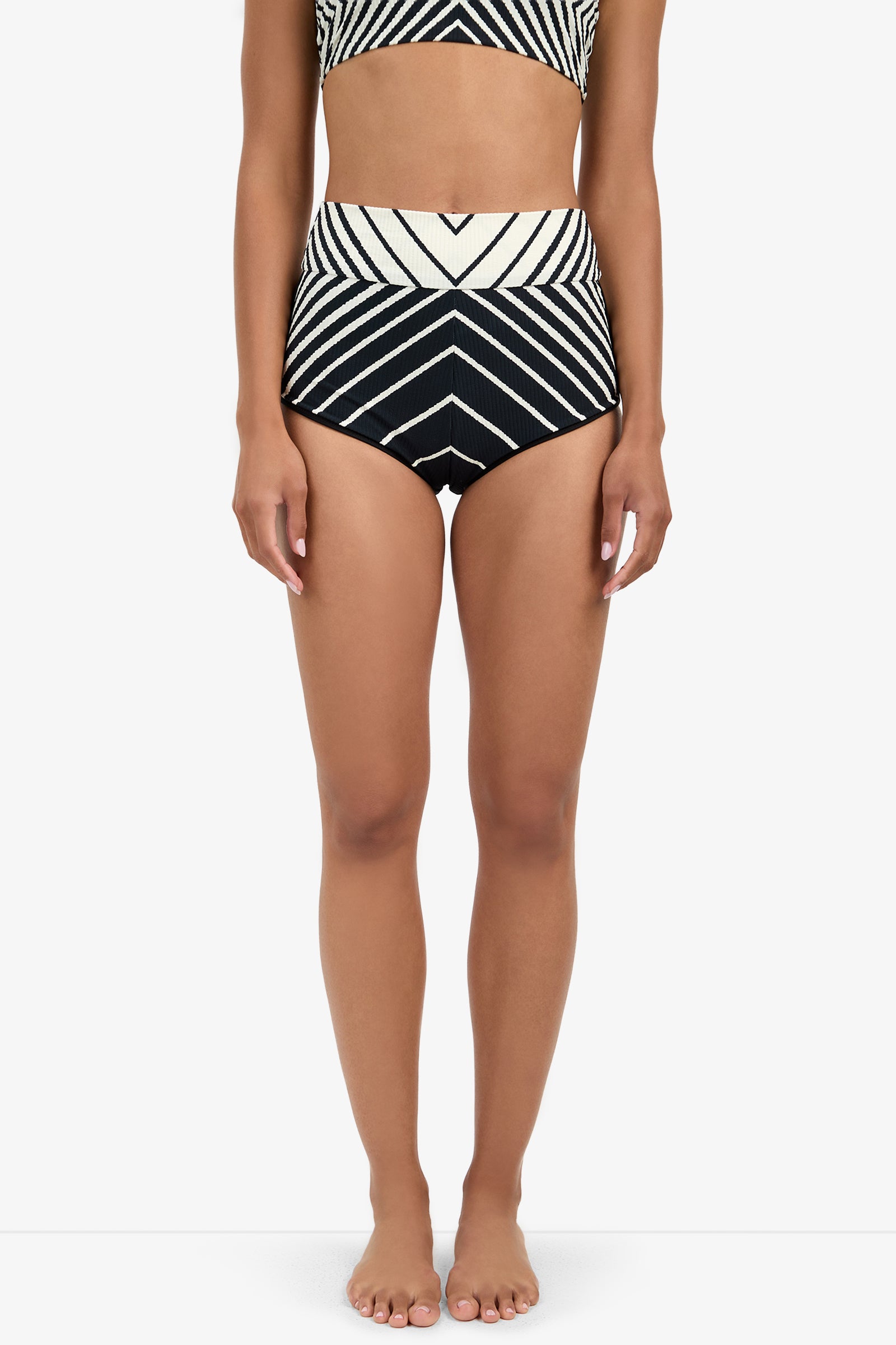 A woman stands barefoot in Net Stripe Swim Shorts, featuring bold black and white high-waisted chevron stripes. The image is cropped from shoulders to feet, set against a plain white background.