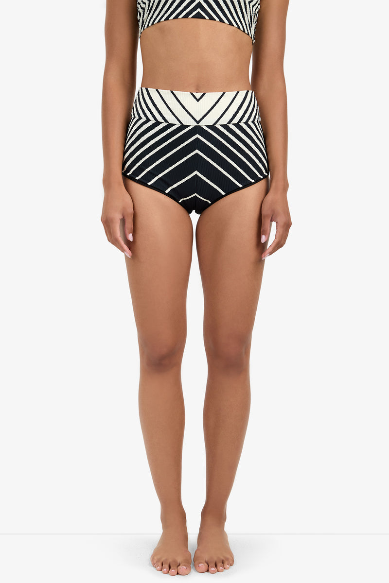 A woman stands barefoot in Net Stripe Swim Shorts, featuring bold black and white high-waisted chevron stripes. The image is cropped from shoulders to feet, set against a plain white background.