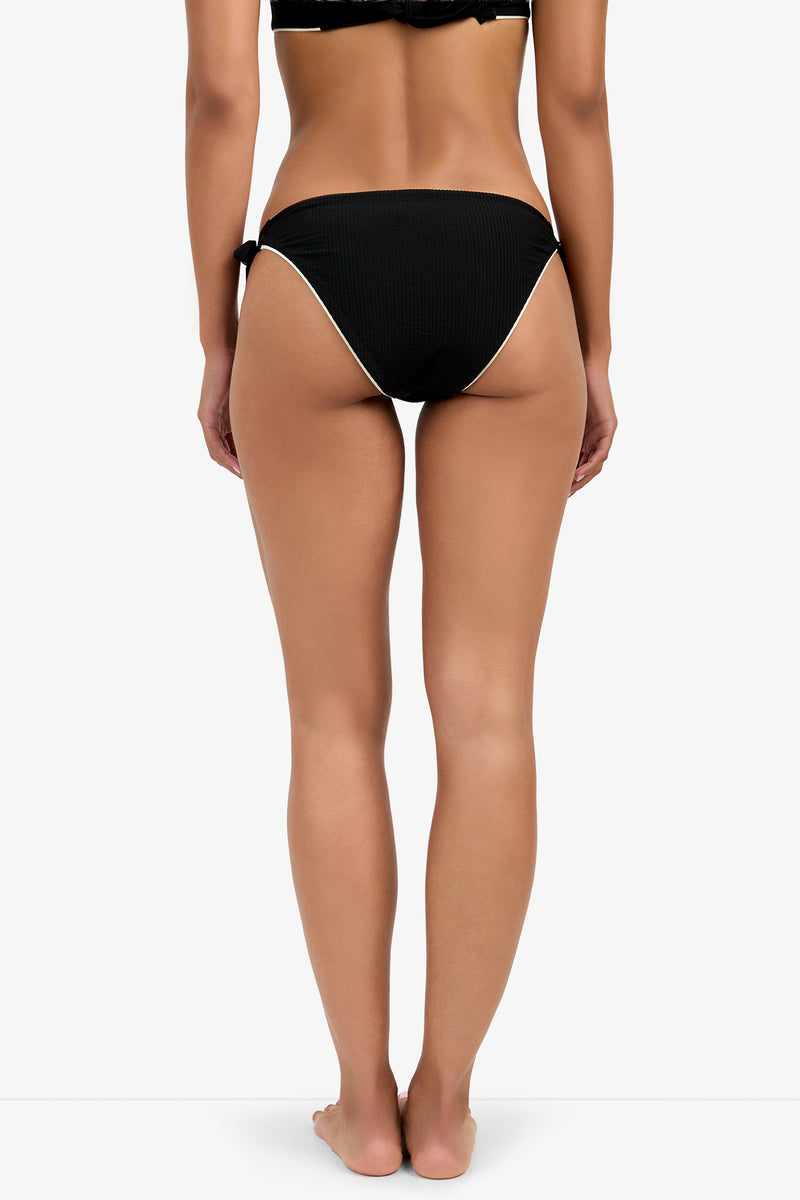 A woman stands barefoot with her back to the camera, modeling the CORINTH TIE SIDE HIPSTER— a black bikini with white trim and side ties—against a plain white background.