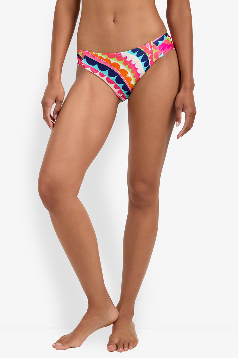 A woman stands barefoot wearing Shirred Hipster bikini bottoms with a scalloped pattern in orange, pink, yellow, blue, and white. The photo is cropped from mid-torso to feet.