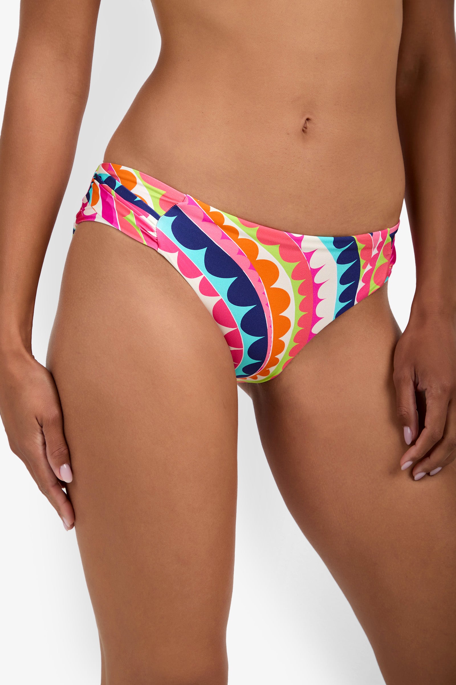 A person wearing Shirred Hipster bikini bottoms with a bold, geometric, scalloped pattern in orange, pink, blue, yellow, and green. The image shows the waist and upper thighs against a white background.