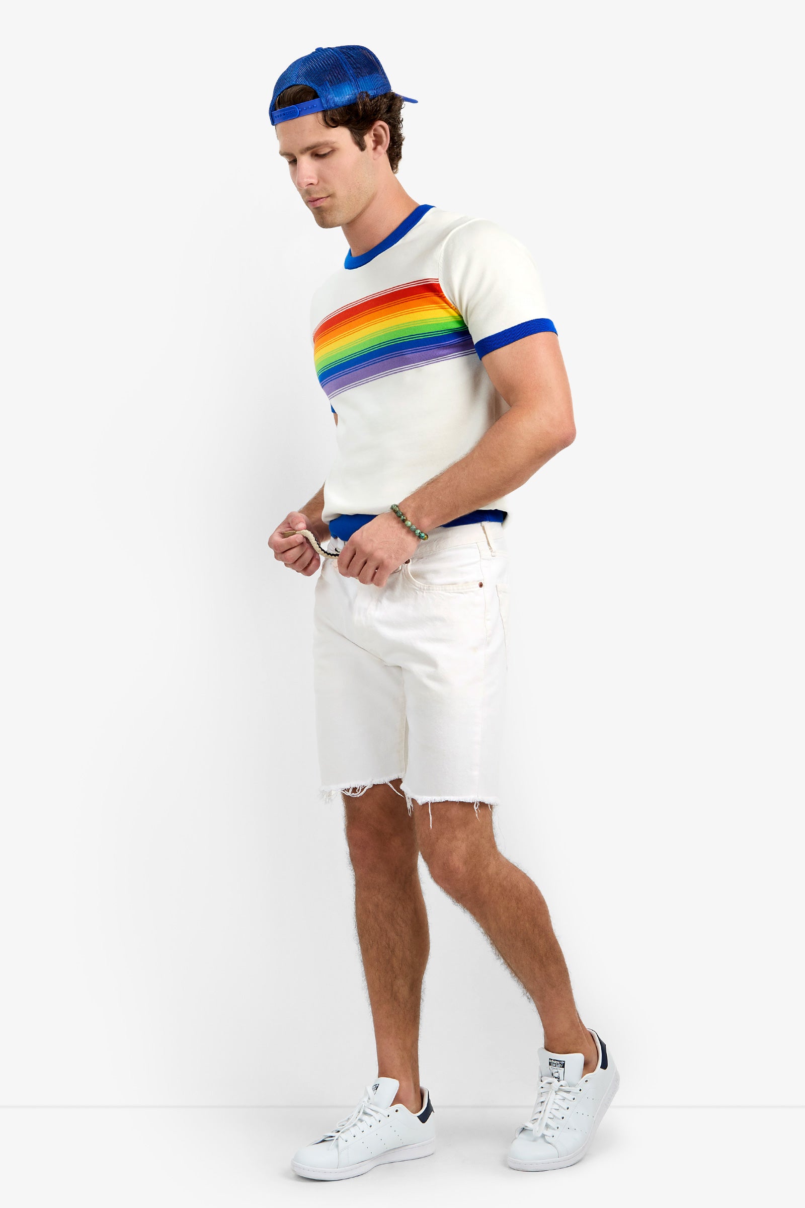 A man wearing the Staley Pride Ringer— a white t-shirt with a rainbow stripe— with a blue cap, white denim shorts, and white sneakers stands against a plain background, looking down and adjusting his shorts.
