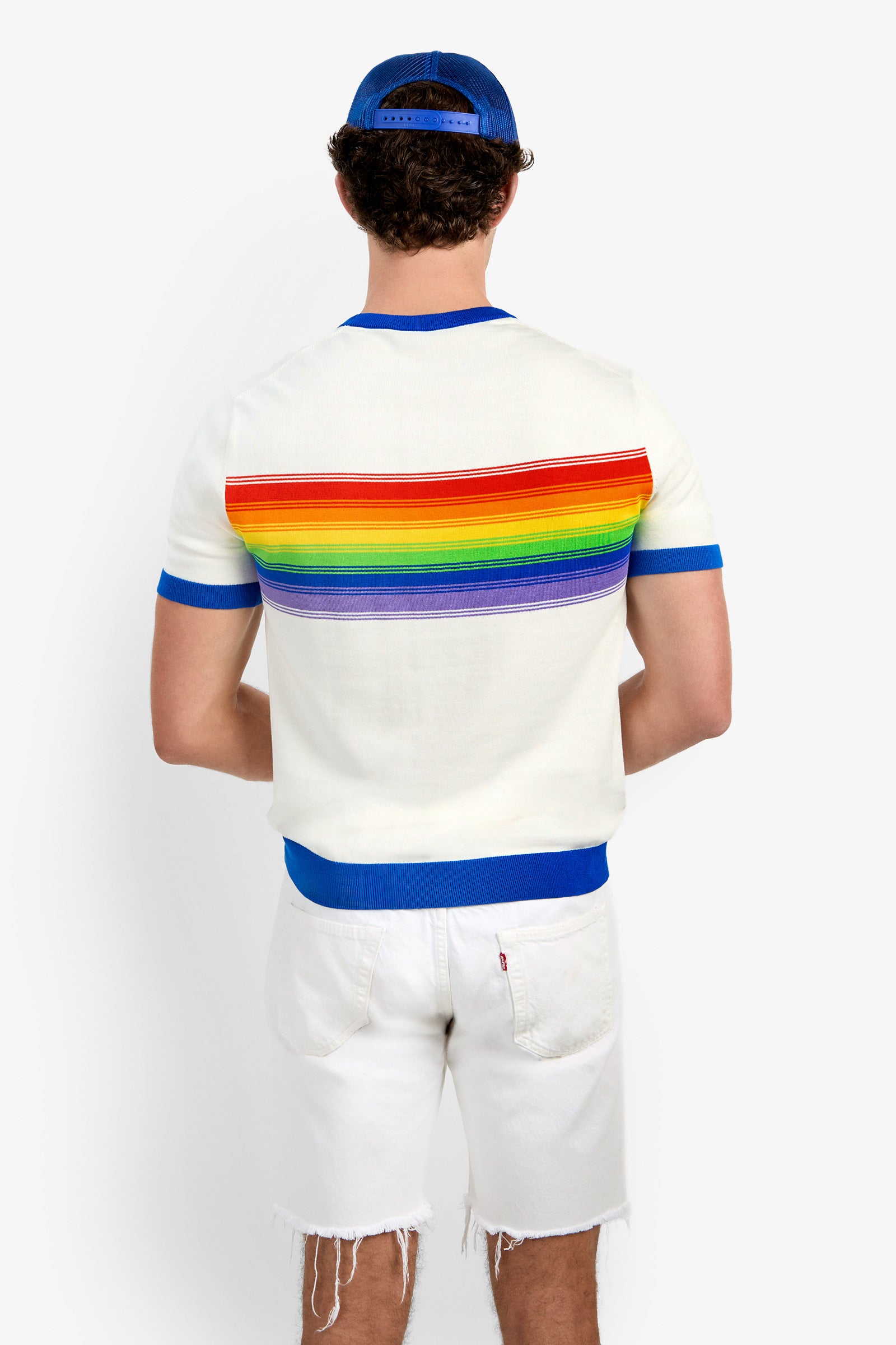 Someone stands facing away from the camera, wearing the Staley Pride Ringer—a white shirt with a rainbow stripe on the back—paired with a blue cap and white frayed shorts, set against a plain background.