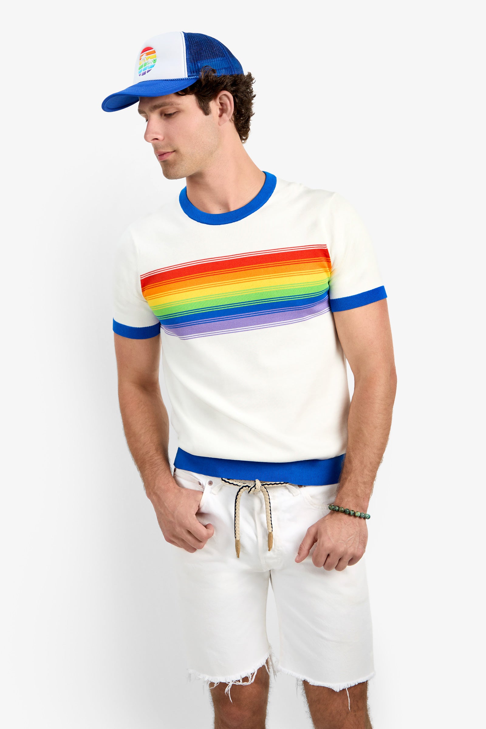 A man wears the Staley Pride Ringer, a white t-shirt with a rainbow stripe and blue trim, paired with white shorts, a braided belt, a blue-and-white mesh cap with a rainbow patch, and a beaded bracelet against a plain background.