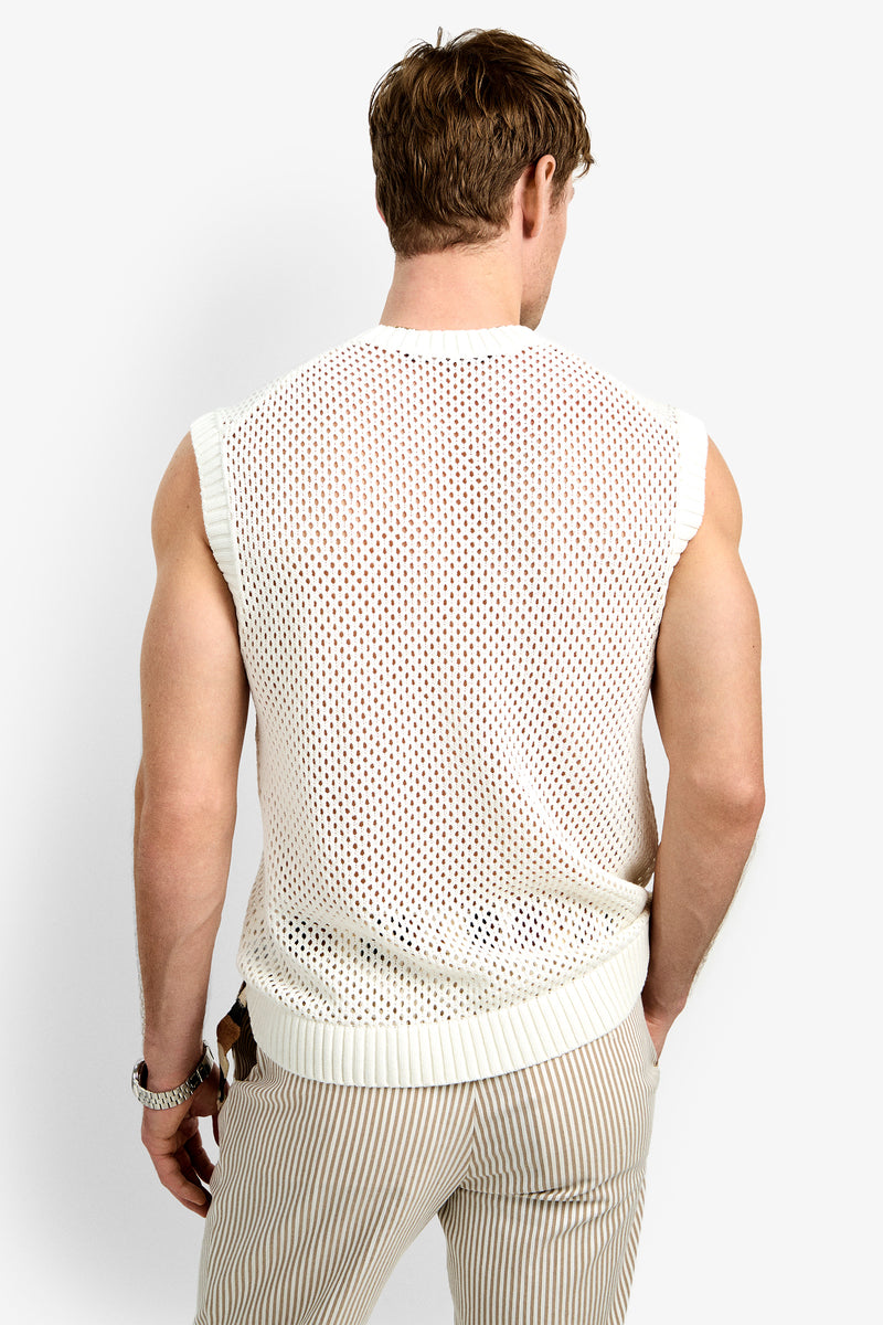 A man faces away from the camera, wearing the KRAMER MESH PULLOVER—a sleeveless white mesh sweater—paired with light striped pants and a bracelet. His right arm rests at his side against a plain white background.