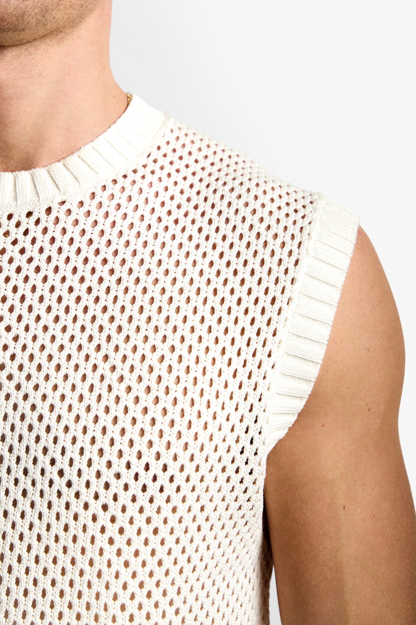 Close-up of a person wearing the KRAMER MESH PULLOVER, a sleeveless white knit top with ribbed neckline and armhole trim, featuring an open-knit mesh pattern for a sheer, net-like look.