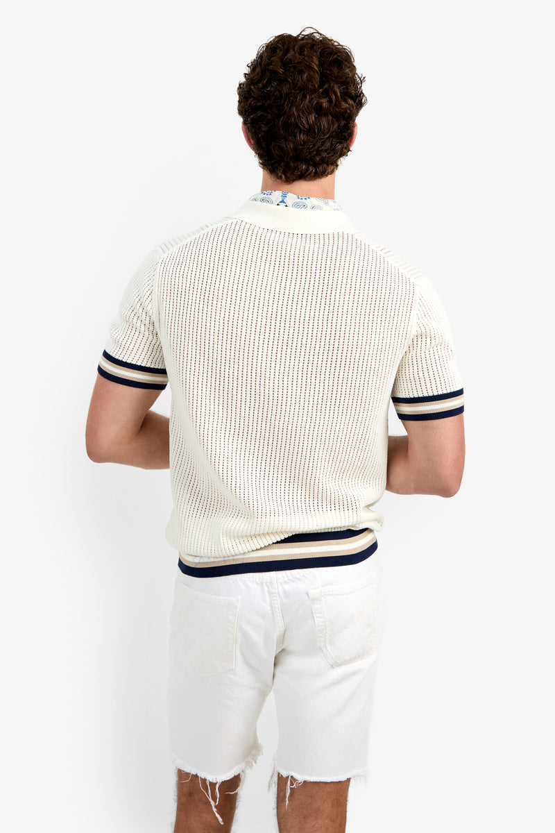 A man with curly brown hair faces away, wearing the Alvin Mesh Polo—a white short-sleeve knit shirt with navy striped sleeves and hem—and white frayed hem shorts, set against a plain white background.