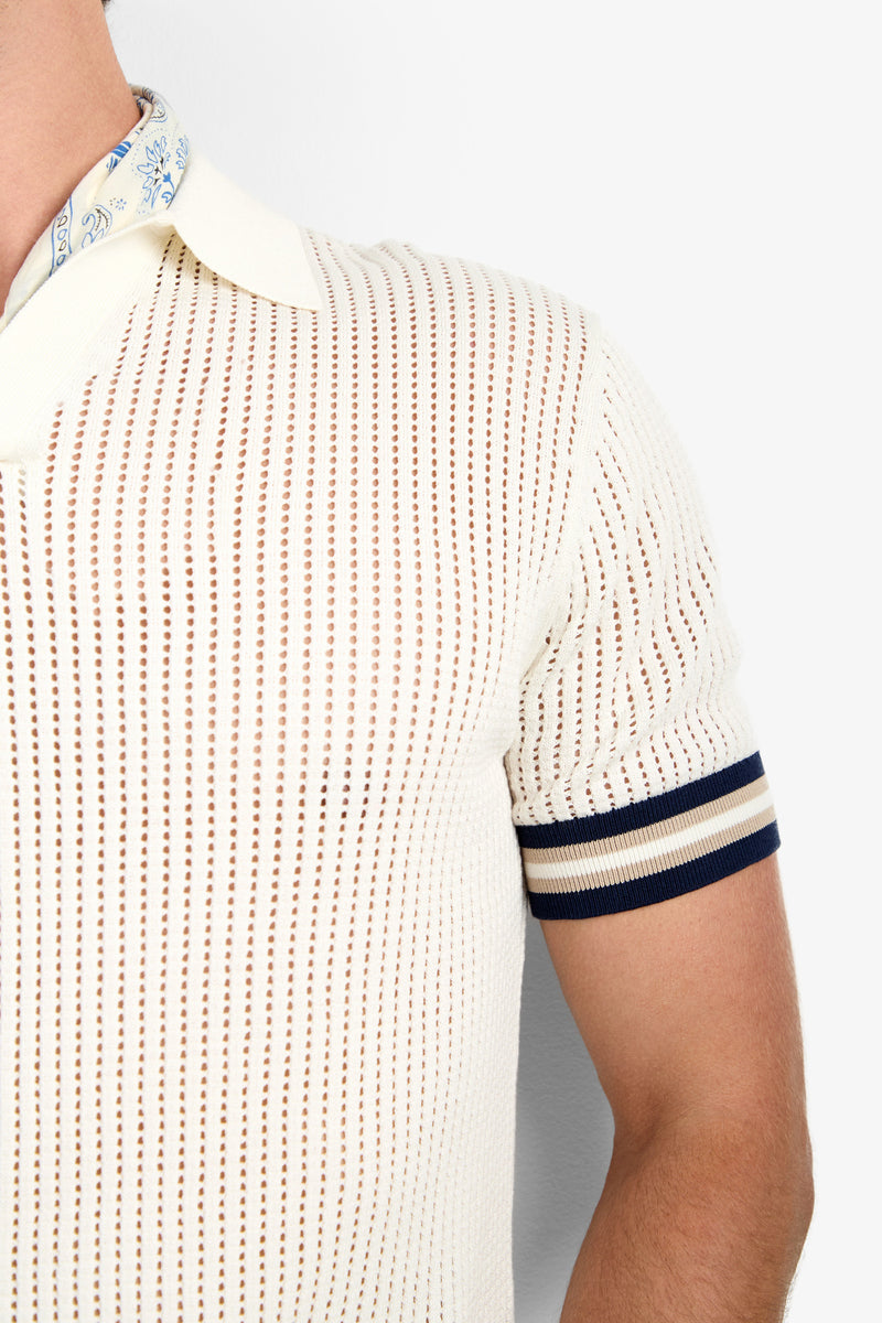 A close-up of a person wearing the Alvin Mesh Polo—a cream short-sleeve knit with vertical mesh stripes, navy-striped cuffs, and a patterned shirt collar visible underneath.
