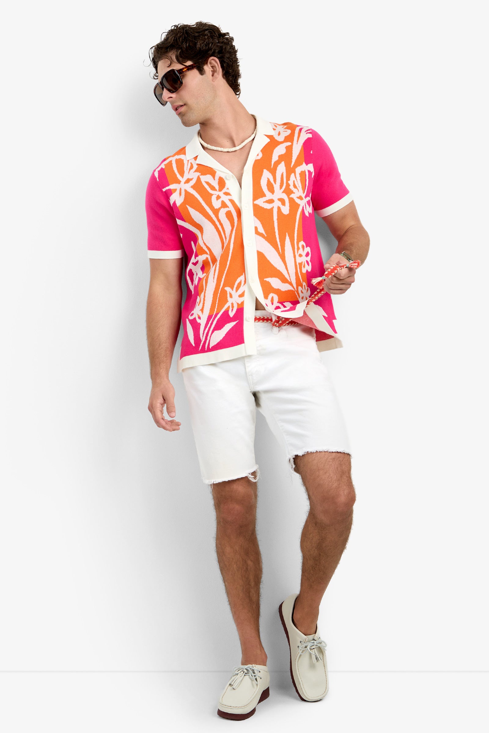 A man wearing the Alessio Polo, a vibrant pink and orange floral shirt, paired with white shorts and sneakers, poses against a white background while holding pink sunglasses and looking to the side.