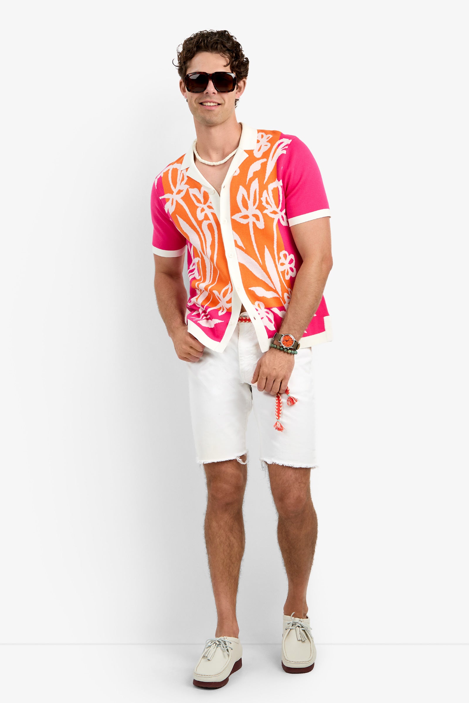 A man wearing sunglasses, a pink and orange floral short-sleeve Alessio Polo, white t-shirt, white ripped shorts, white shoes, and accessories stands smiling against a plain white background.