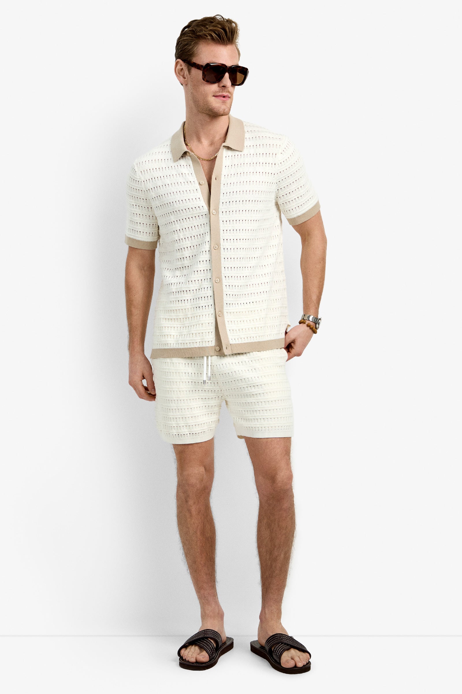 A man wears the Hillcrest Resort Polo, a cream cotton short-sleeve knit shirt over a beige undershirt, with matching knit shorts, sunglasses, a watch, and black slide sandals, standing against a plain white background.
