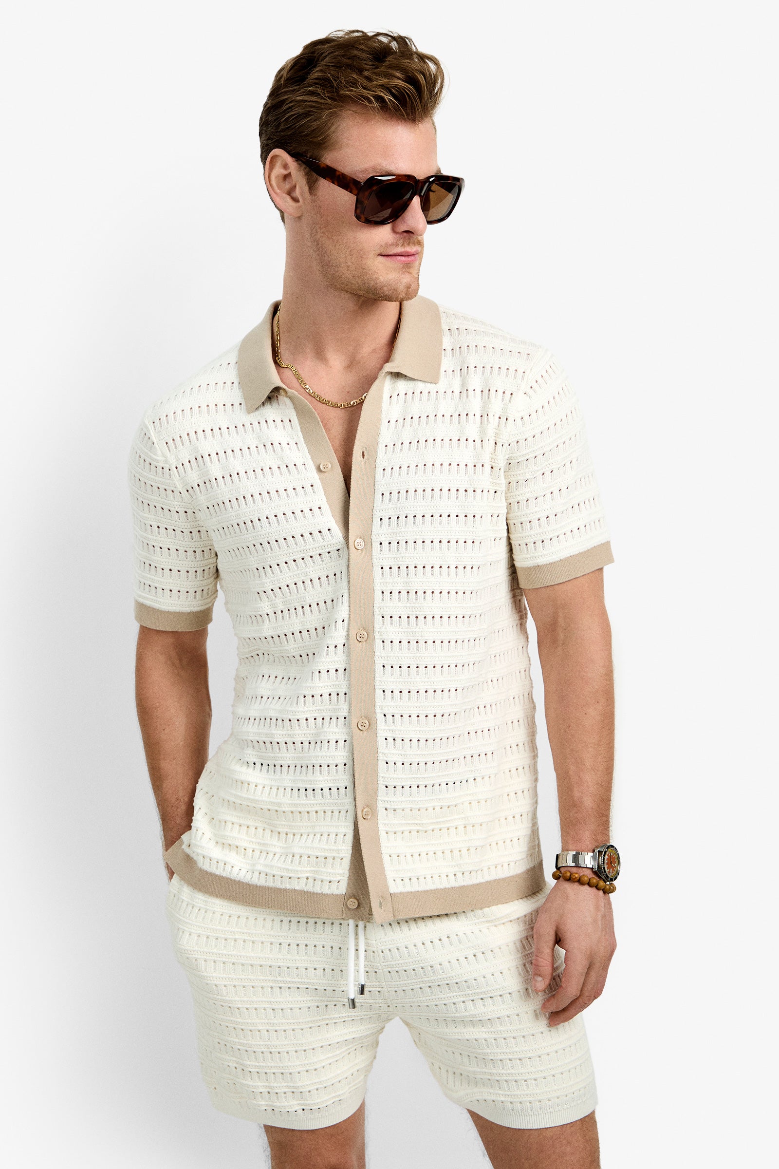 A man in the Hillcrest Resort Polo—short-sleeved, cream with tan trim—and matching 100% cotton shorts stands hands-in-pockets against a plain white background, wearing sunglasses.