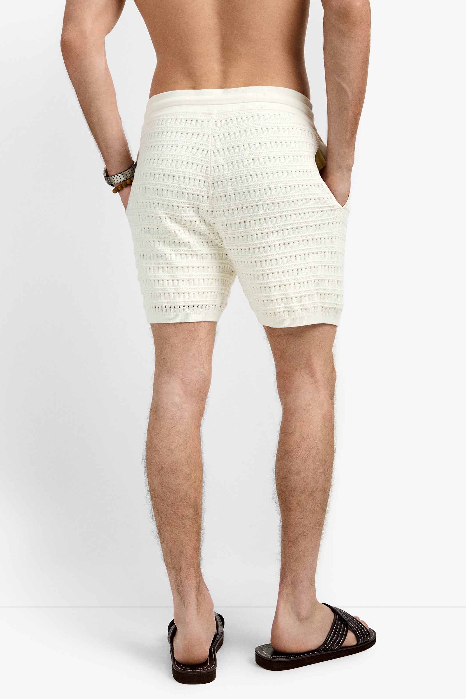 A man stands against a plain white background, hands in his pockets, wearing textured cream-colored Julio Crochet Shorts, a beaded bracelet, and dark brown slide sandals.