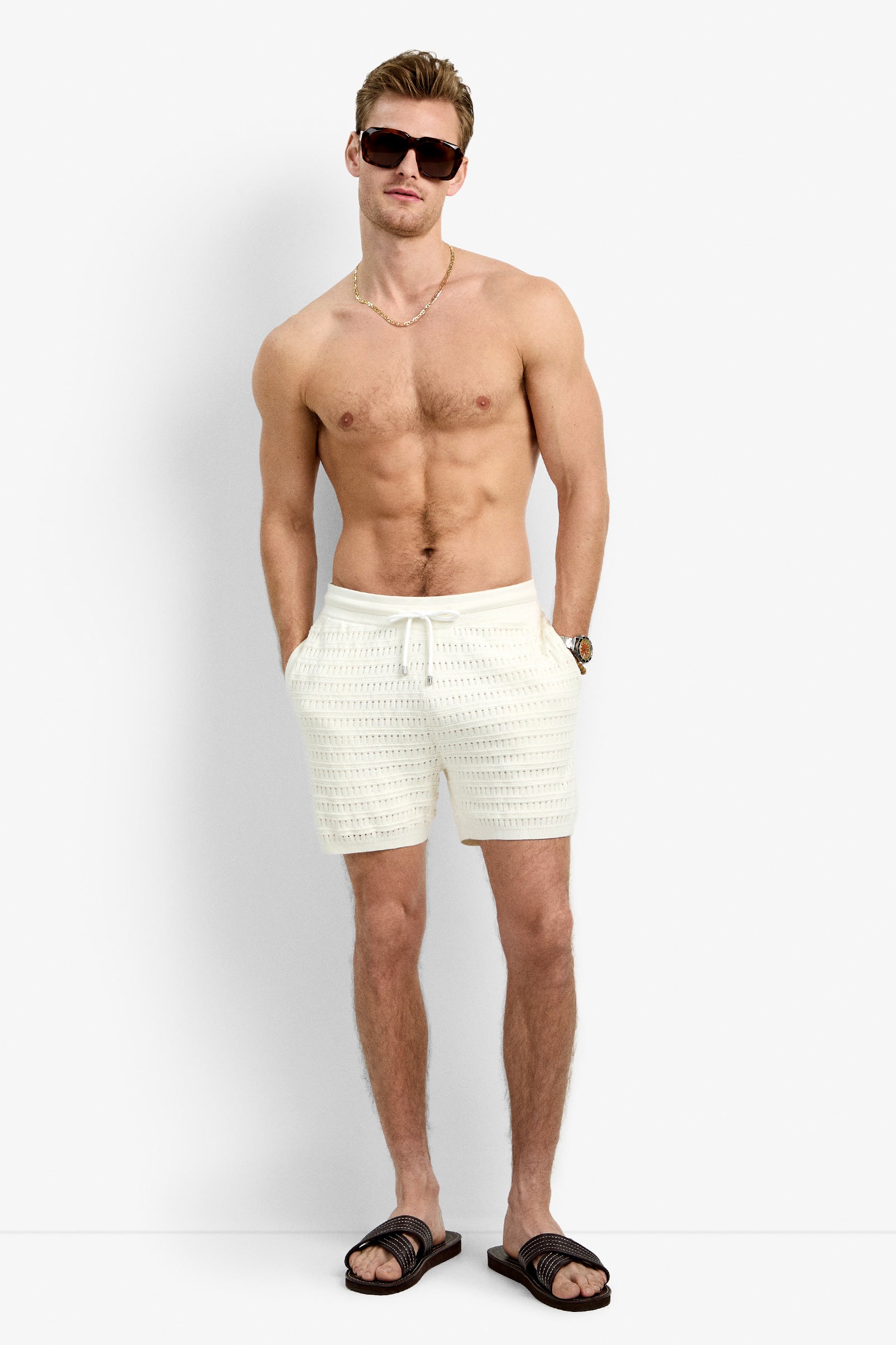 A shirtless man wearing the Julio Crochet Short, black sandals, sunglasses, a gold necklace, and a watch stands against a plain white background with his hands in his pockets.