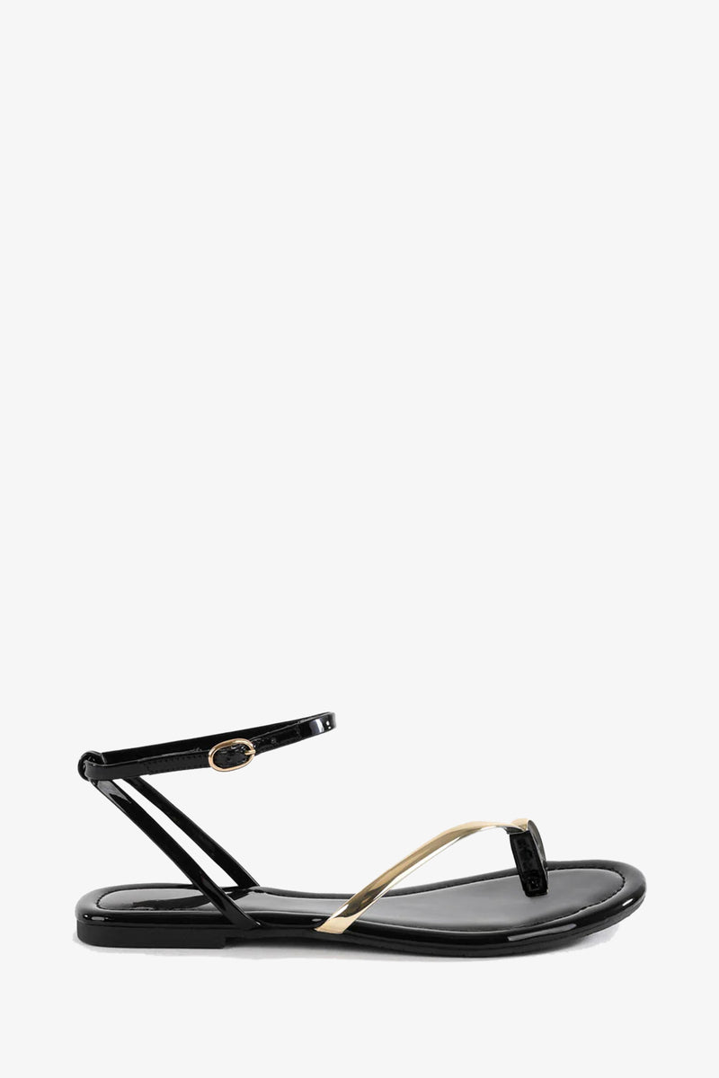 The A Little Extra Sandal is a sleek black flat with a thin ankle strap, simple thong design, and metallic gold accent on the front strap, shown against a white background.
