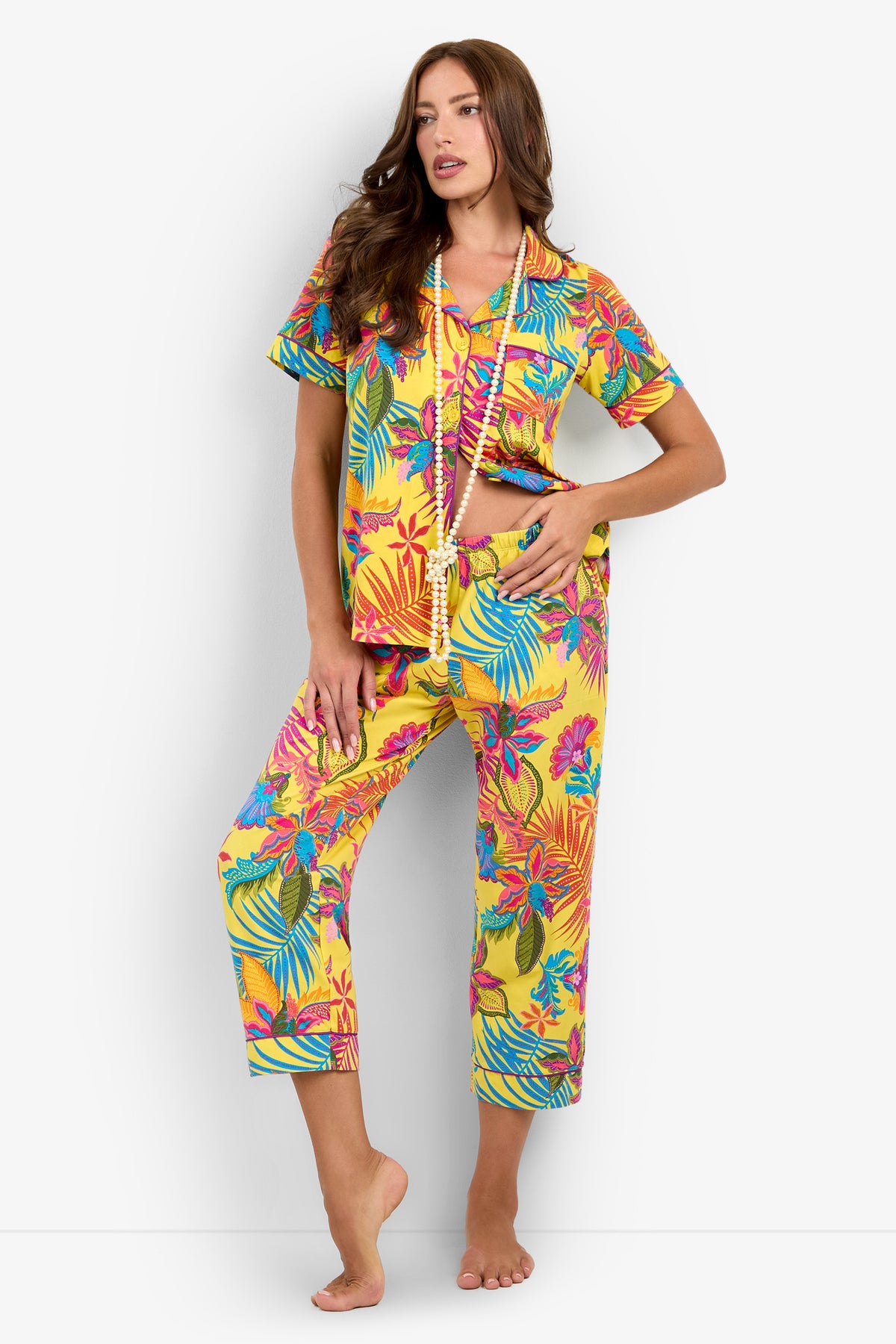 A woman stands barefoot against a plain white background, wearing the Jacobean Short Sleeve Cropped PJ Set in a vibrant yellow tropical print and accessorized with two long beaded necklaces.