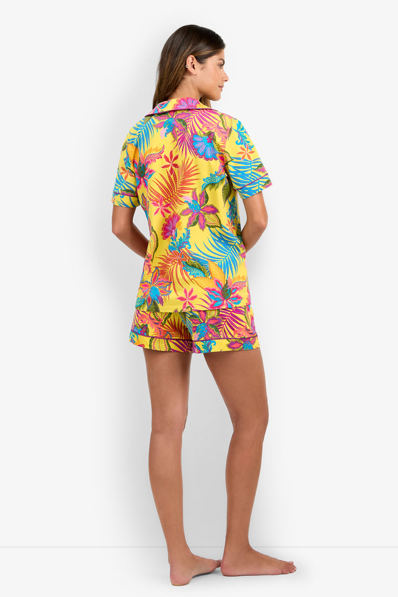 A woman stands barefoot facing away, wearing the Jacobean Short Sleeve PJ Set—a yellow pajama ensemble featuring vibrant tropical floral patterns, short sleeves, and matching shorts—set against a plain white background.