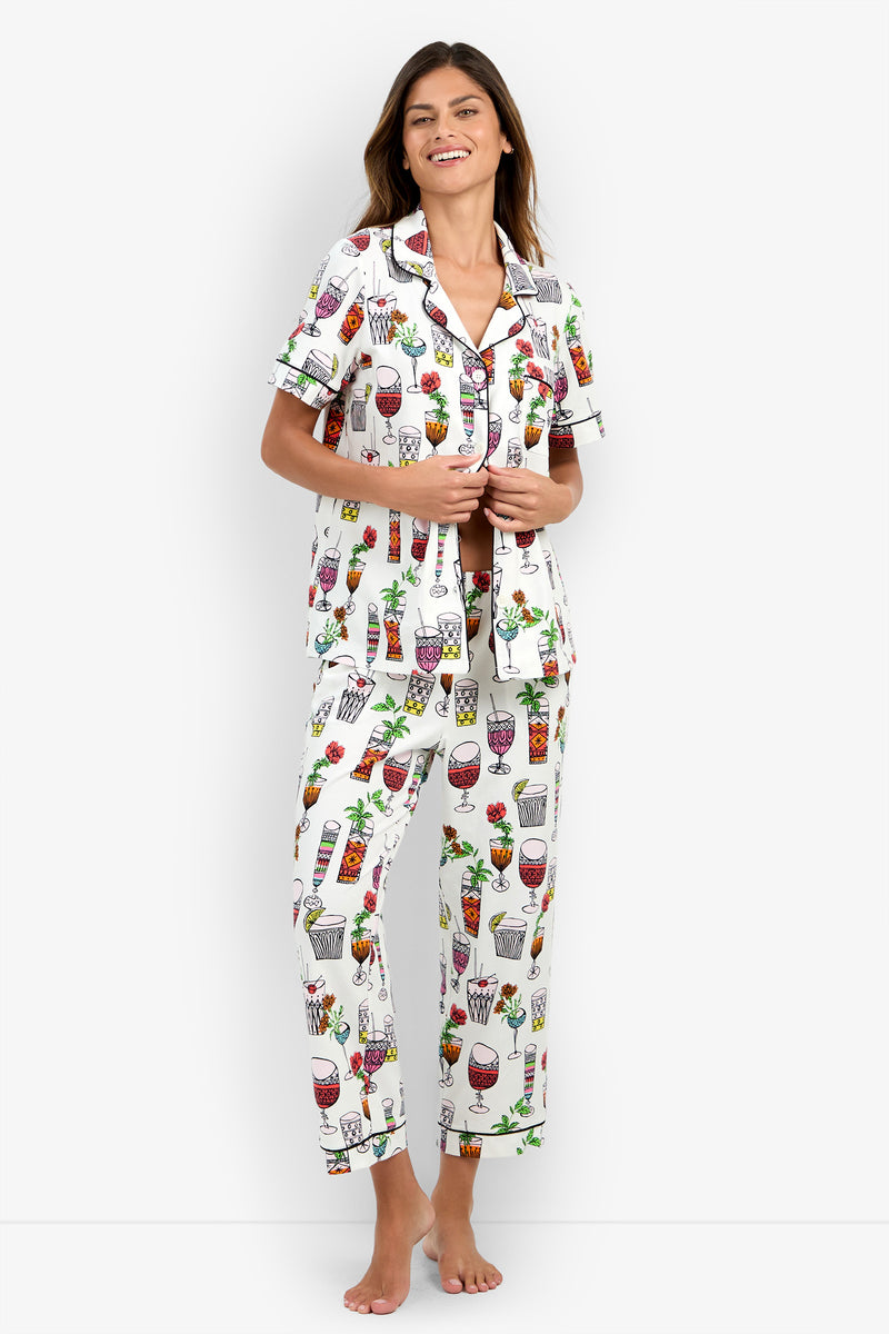 A woman stands and smiles, wearing the Short Sleeve Cropped Pajamas Set featuring a white base with colorful drink and cocktail glass prints, a short-sleeve button-up top, and matching long pants. The photo is taken against a plain, light-colored studio background.