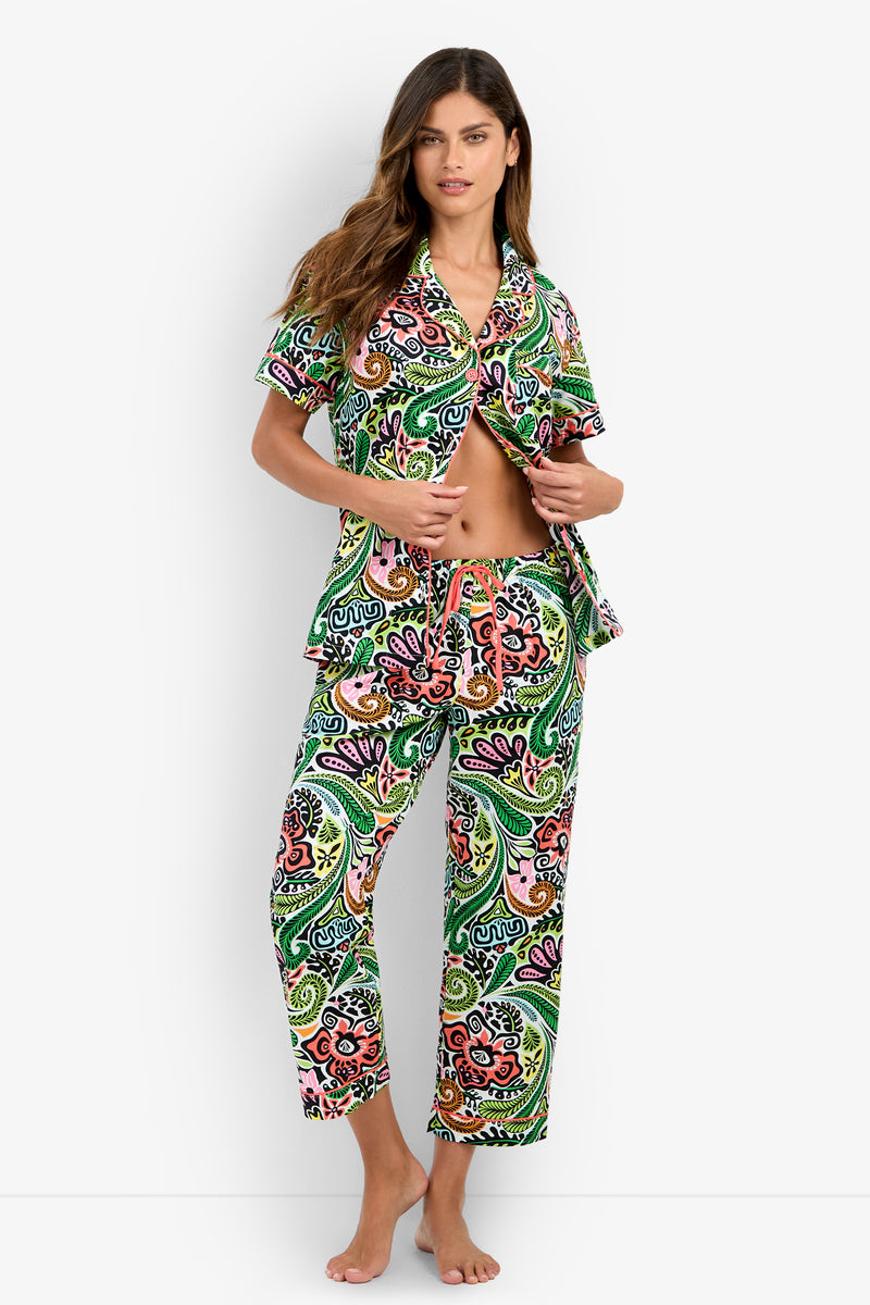 A woman stands barefoot against a plain white background, wearing the Short Sleeve Cropped Pajamas Set featuring a colorful paisley pattern, holding the shirt open to reveal her midriff.