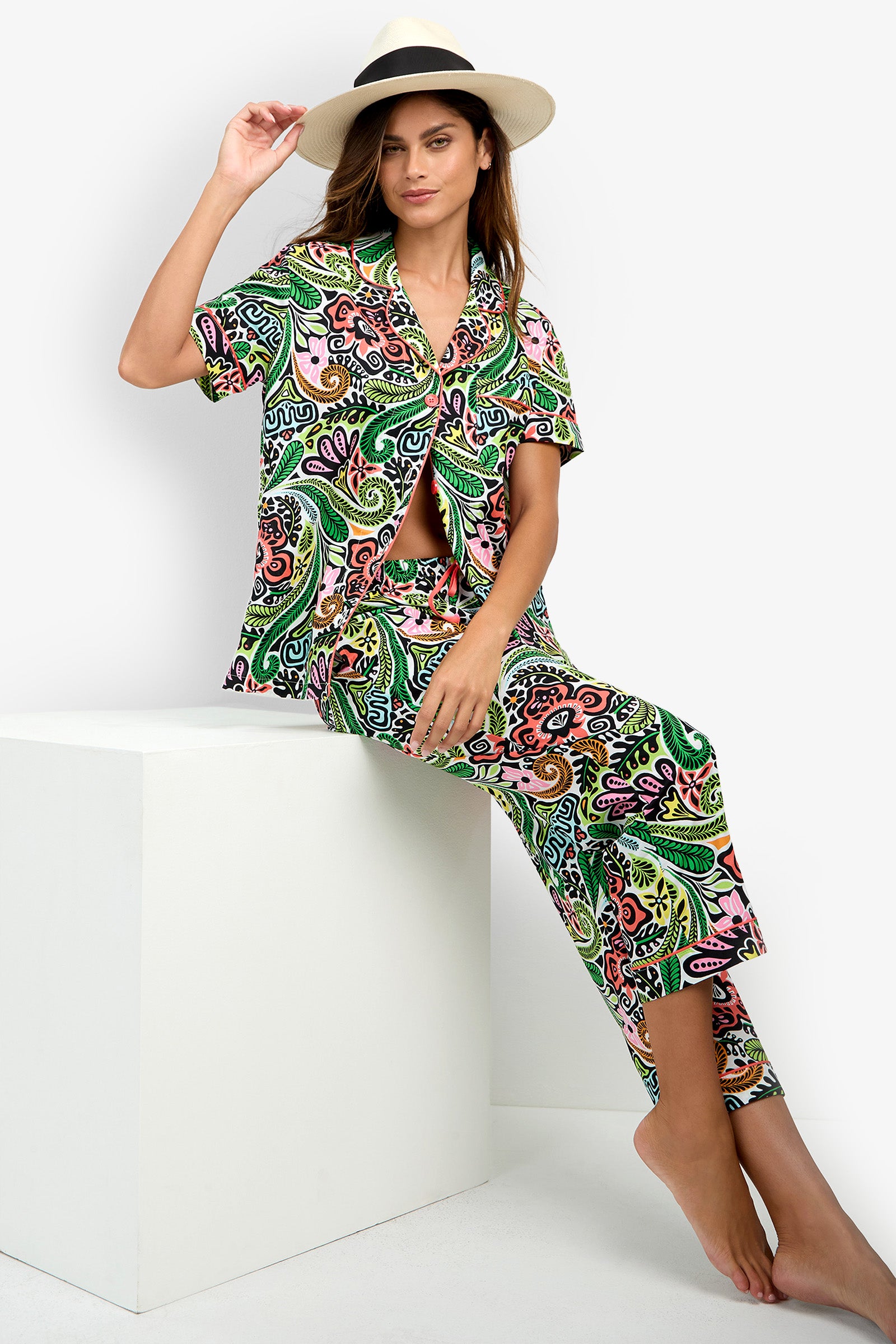 A woman wearing the Short Sleeve Cropped Pajamas Set in a vibrant pattern and a wide-brim hat sits barefoot on a white cube against a plain white background, gazing at the camera while lifting the brim of her hat with one hand.