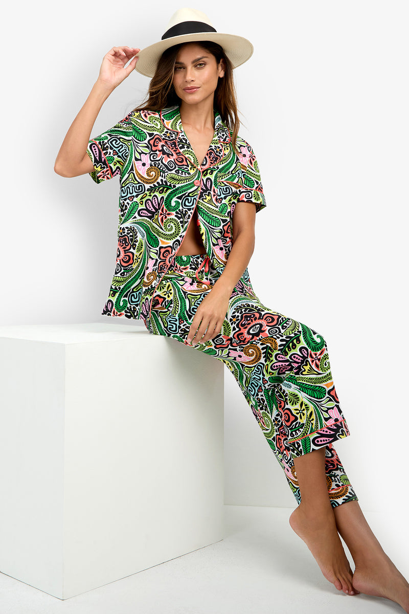 A woman wearing the Short Sleeve Cropped Pajamas Set in a vibrant pattern and a wide-brim hat sits barefoot on a white cube against a plain white background, gazing at the camera while lifting the brim of her hat with one hand.