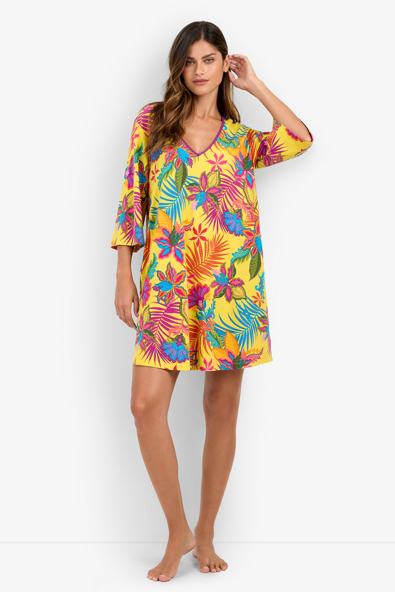 A woman poses barefoot against a white background wearing a loose, knee-length yellow sleepshirt featuring pink, blue, and green tropical floral and leaf patterns.