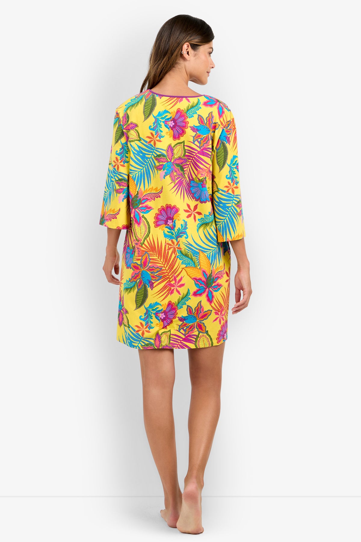A woman stands barefoot facing away, wearing a loose-fitting, knee-length sleepshirt in yellow with bright, colorful tropical floral patterns.