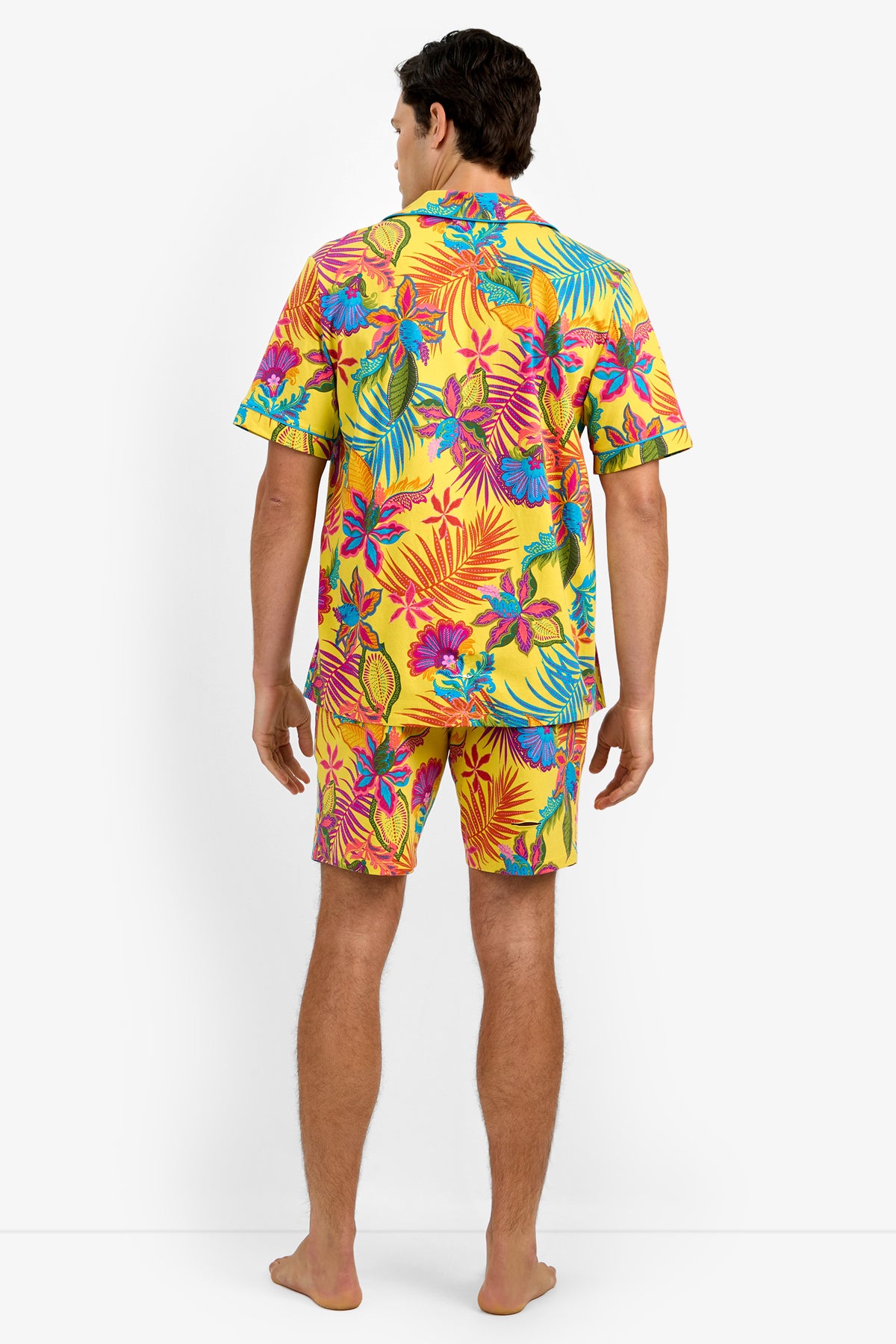 A man stands barefoot facing away, wearing Short Sleeve Boxer Pajamas featuring a matching yellow shirt and shorts with a vibrant tropical floral pattern of pink, blue, and orange leaves and flowers.