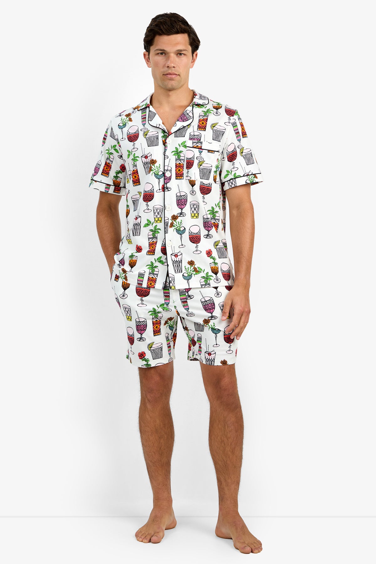 A man stands barefoot wearing Short Sleeve Boxer Pajamas in Happy Hour, featuring a colorful cocktail print. He poses with one hand in his pocket against a plain white background.