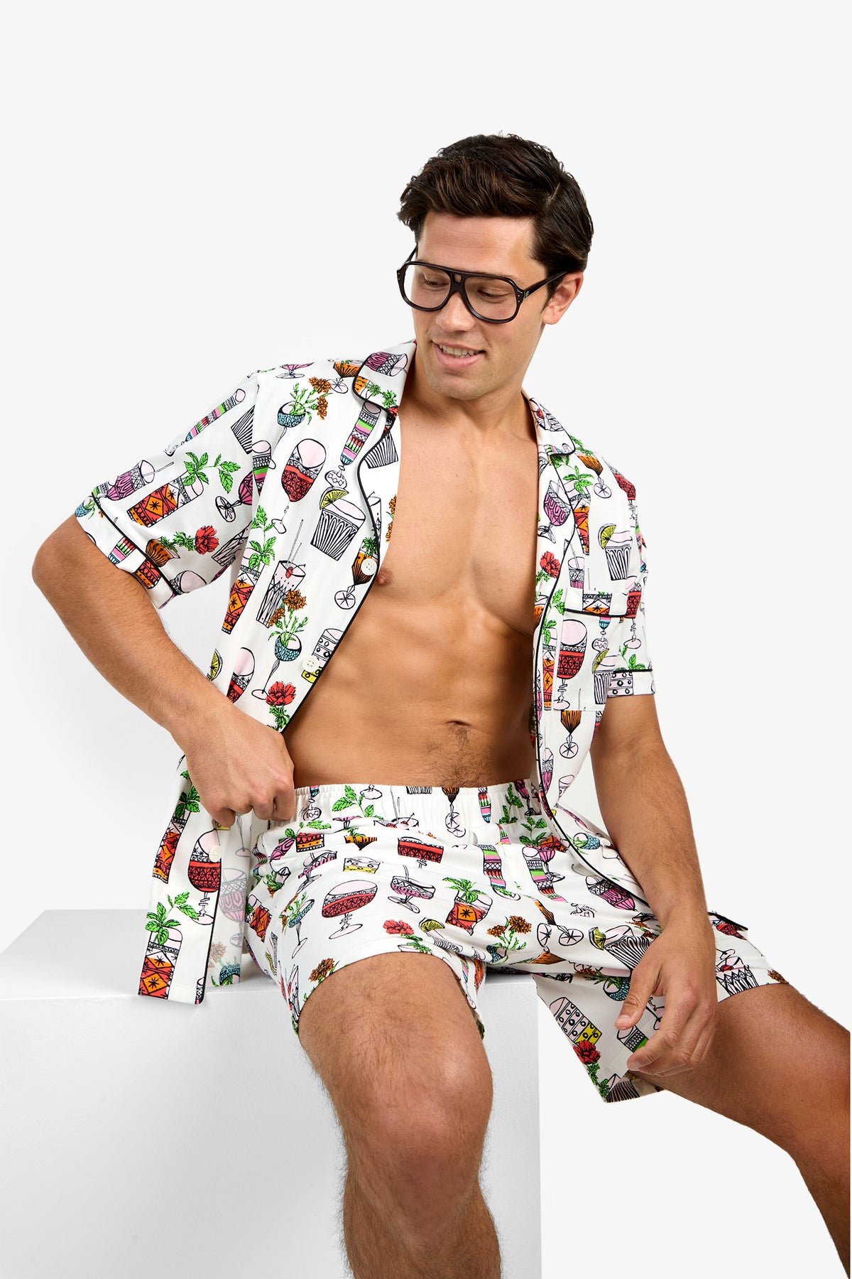 A man with dark hair and glasses sits on a white block, wearing Short Sleeve Boxer Pajamas featuring colorful cocktail and drink prints, his shirt open to reveal his bare chest.